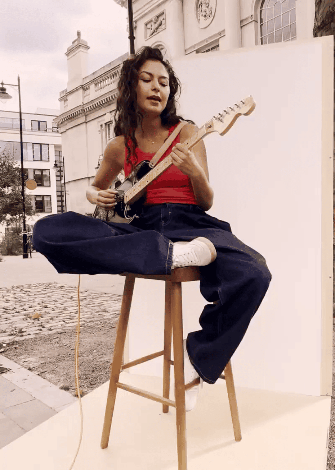 Woman in a red top and jeans plays an electric guitar while seated on a stool outdoors, with buildings in the background.