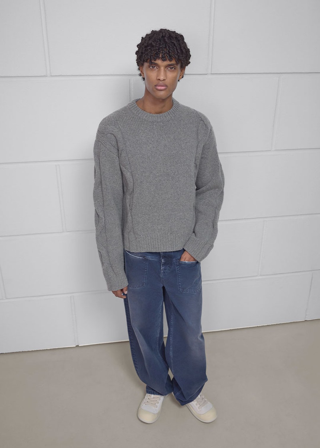 Person standing against a gray wall wearing a thick gray sweater, loose-fitting blue jeans, and white sneakers.