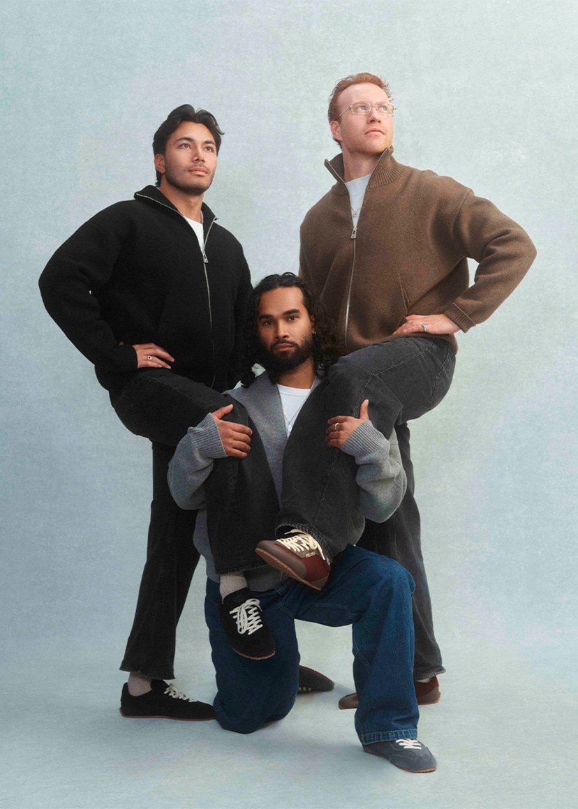 Three men pose confidently in layered outfits against a light background. Two stand, one kneels; all have serious expressions.