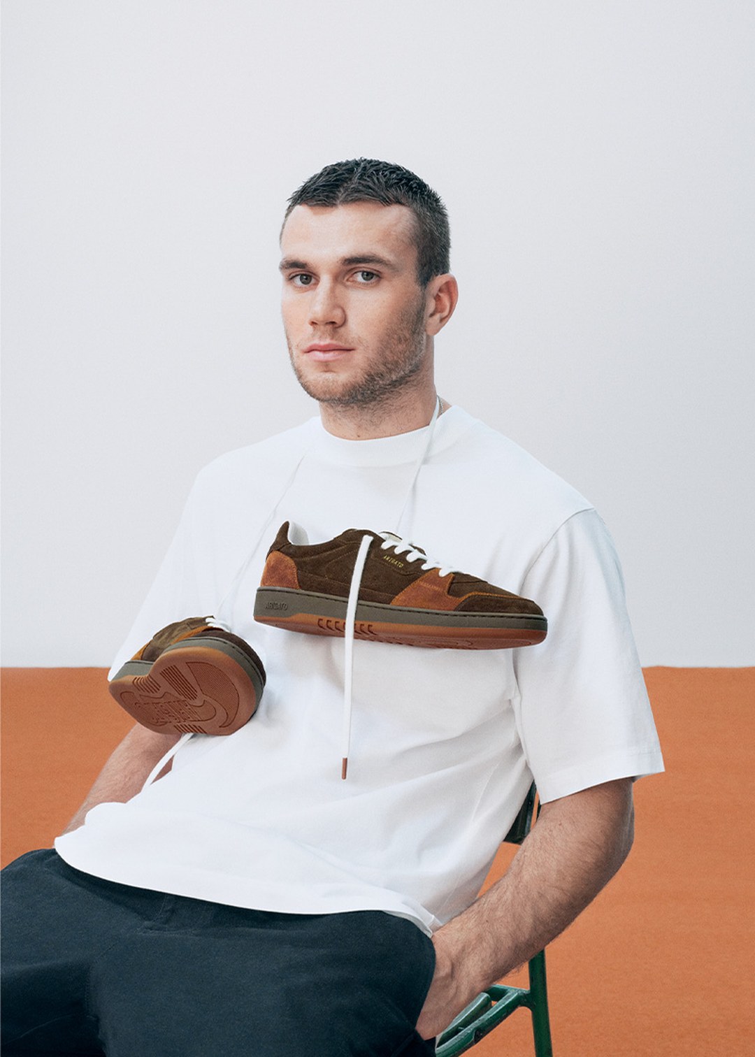 A man in a white t-shirt sits on a chair with brown sneakers draped around his neck, against a white and orange background.