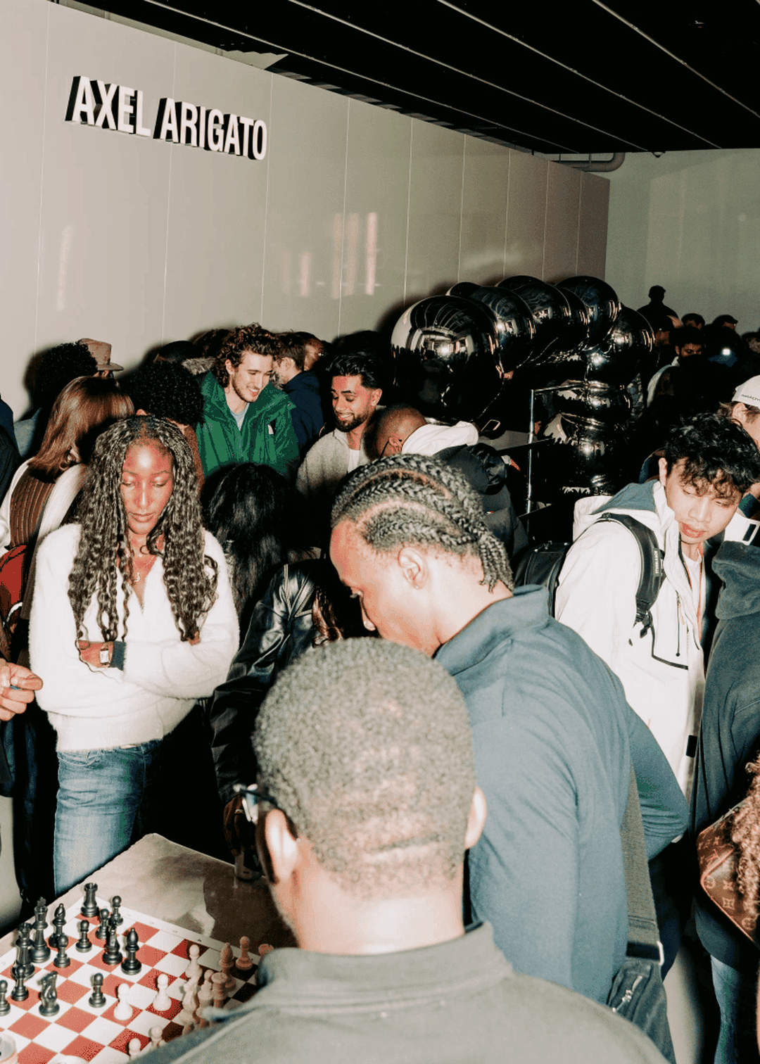 Crowd gathered around a chess game at an Axel Arigato event; people watching and engaging in conversation in a dimly lit space.