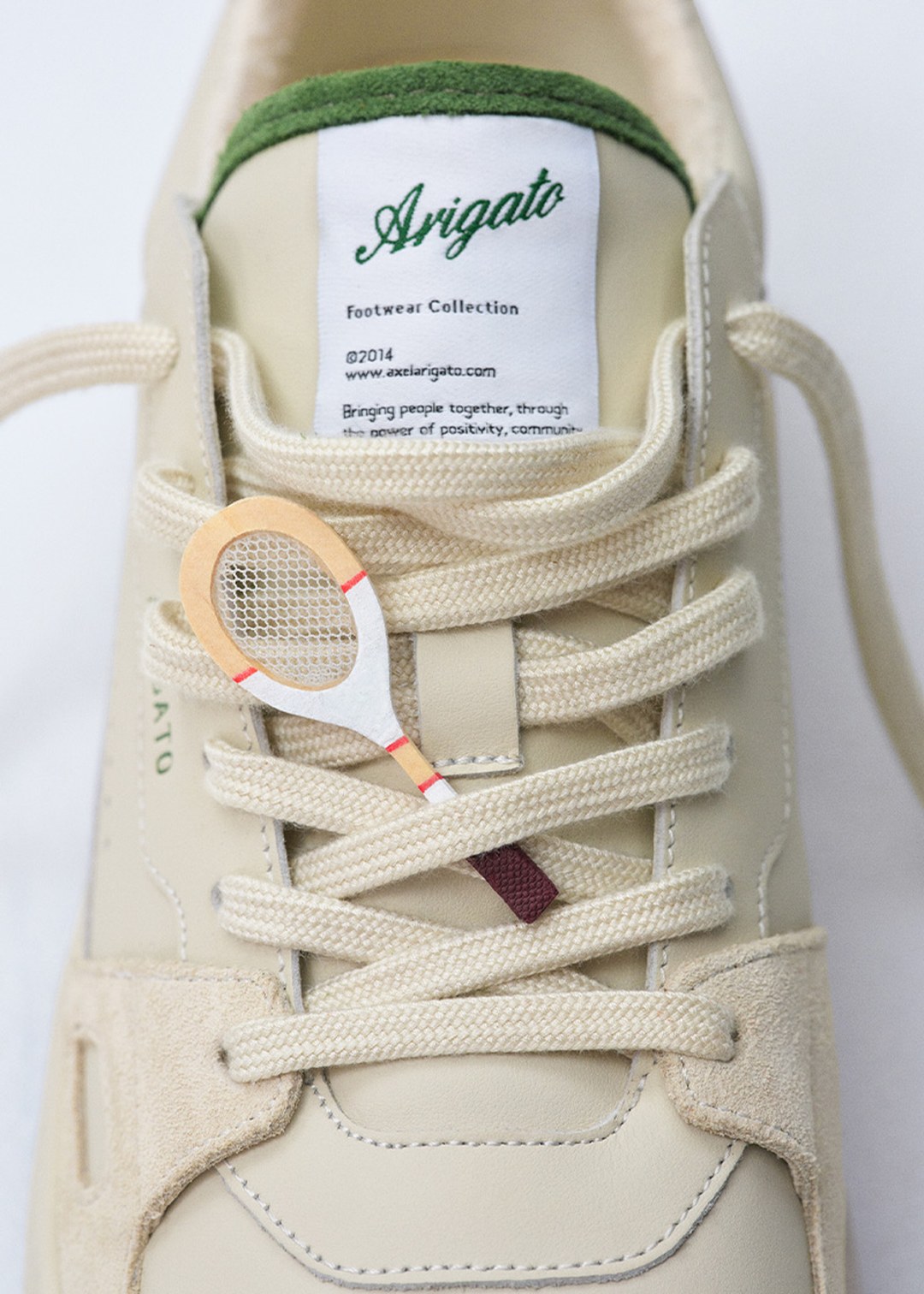 Close-up of a beige sneaker with cream laces, featuring a mini tennis racket charm. "Arigato" label is visible at the top of the shoe tongue.