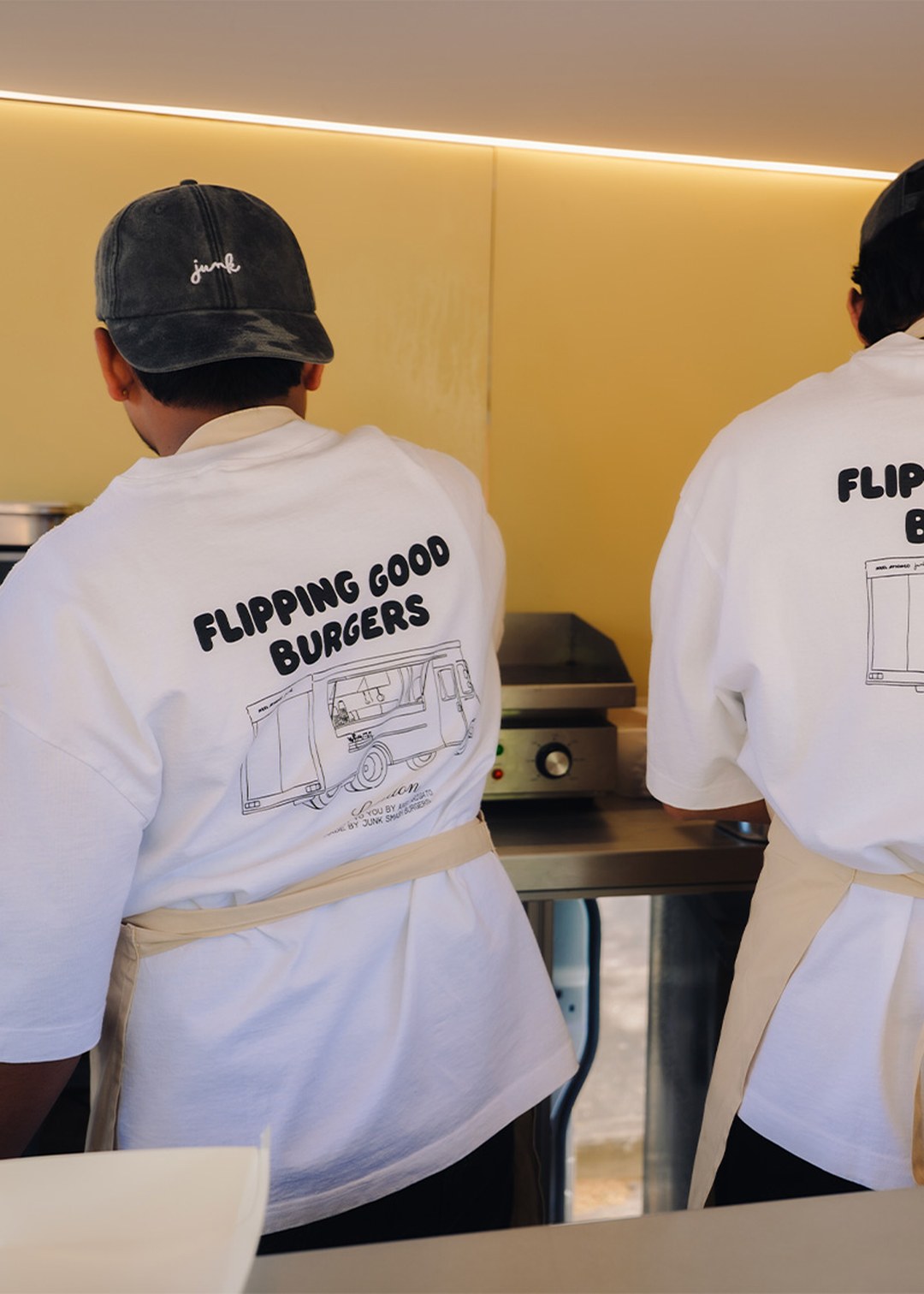 Two people in white shirts and aprons work in a food truck, with "Flipping Good Burgers" printed on their backs.