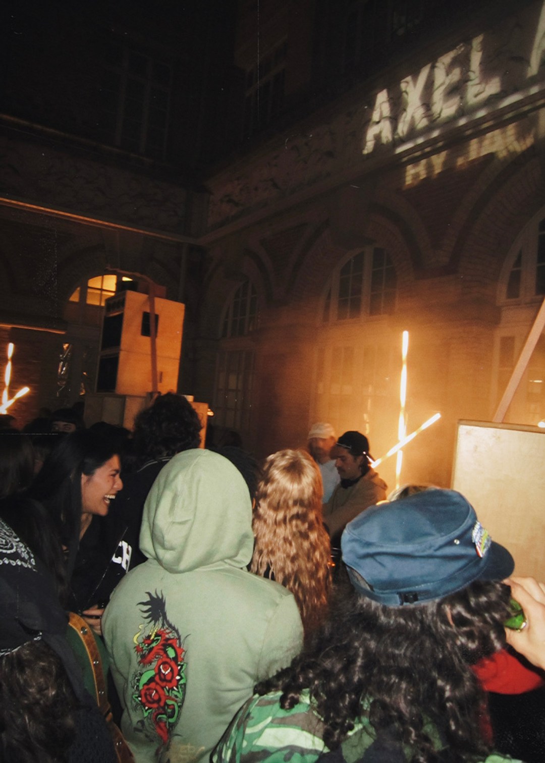 Group of people gathers, some smiling, in a dimly lit outdoor area with graffiti overhead and sparklers illuminating the scene.