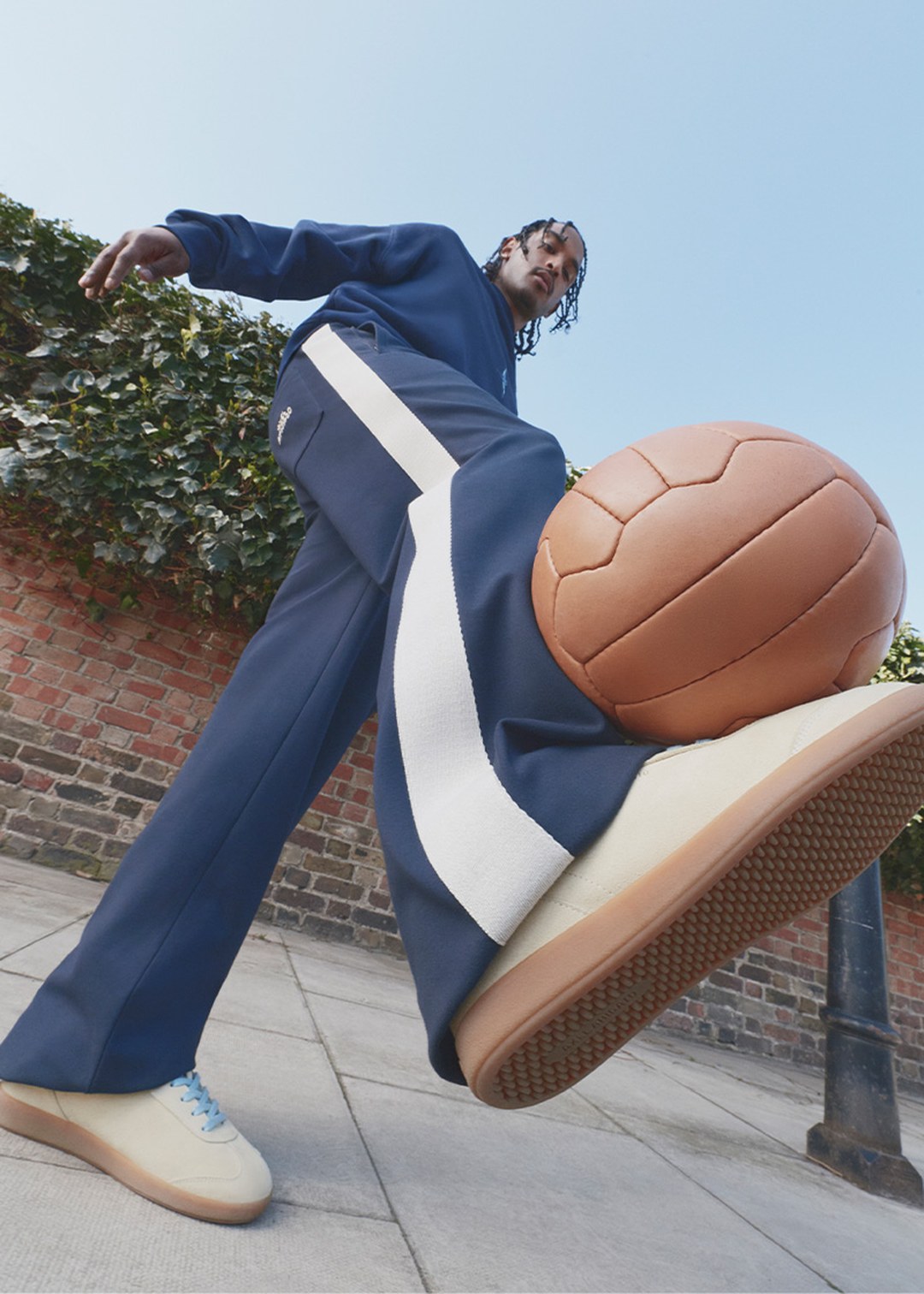 Person in blue tracksuit and sneakers, balancing a vintage-style leather football on the sidewalk, with a brick wall and greenery in the background.
