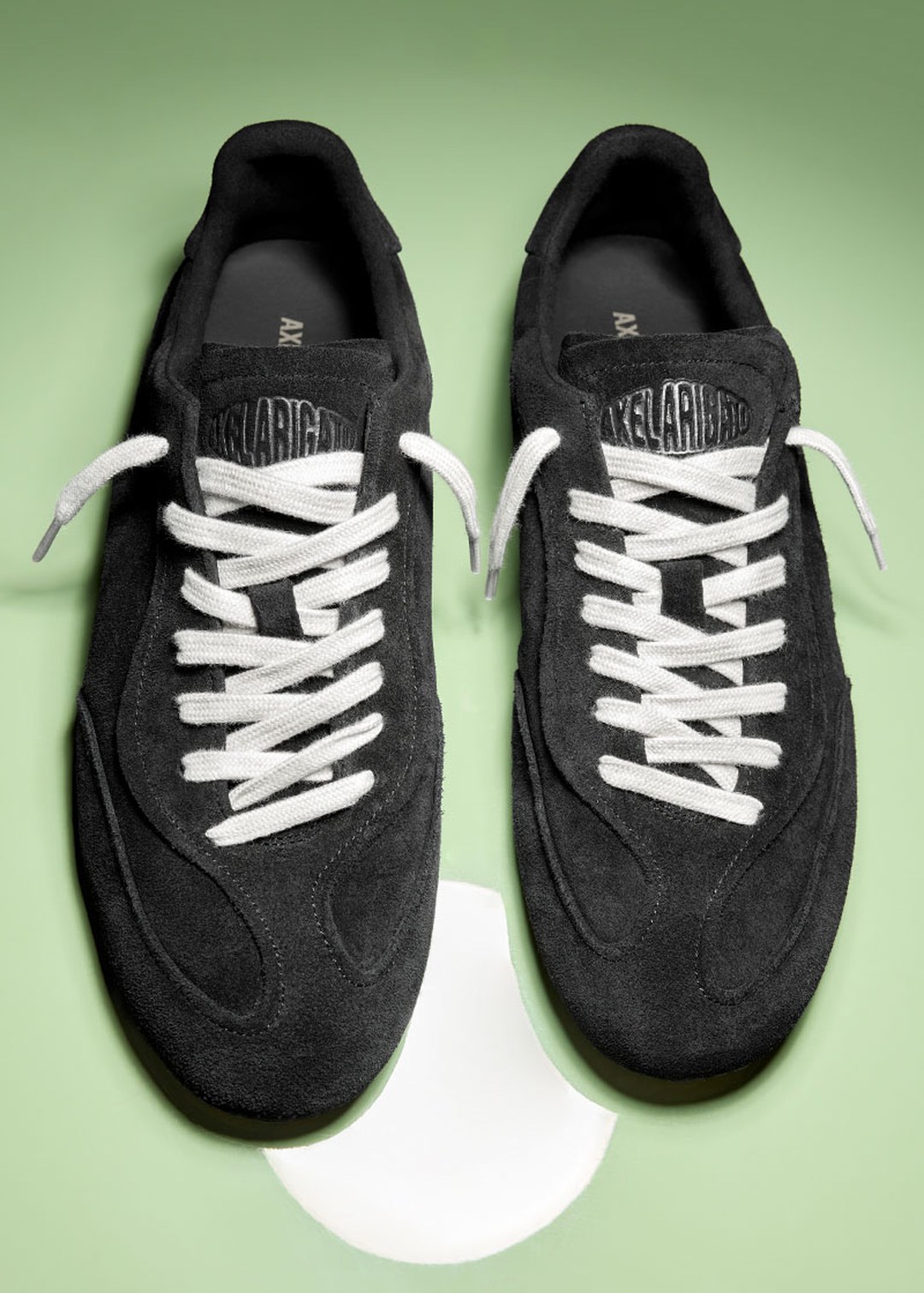 Black suede sneakers with white laces on a light green background, featuring a circular white spot beneath the shoes.