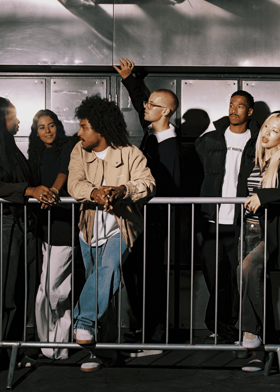 A group of six young adults casually pose around a metal barrier, some leaning and others standing, against a metallic urban backdrop.