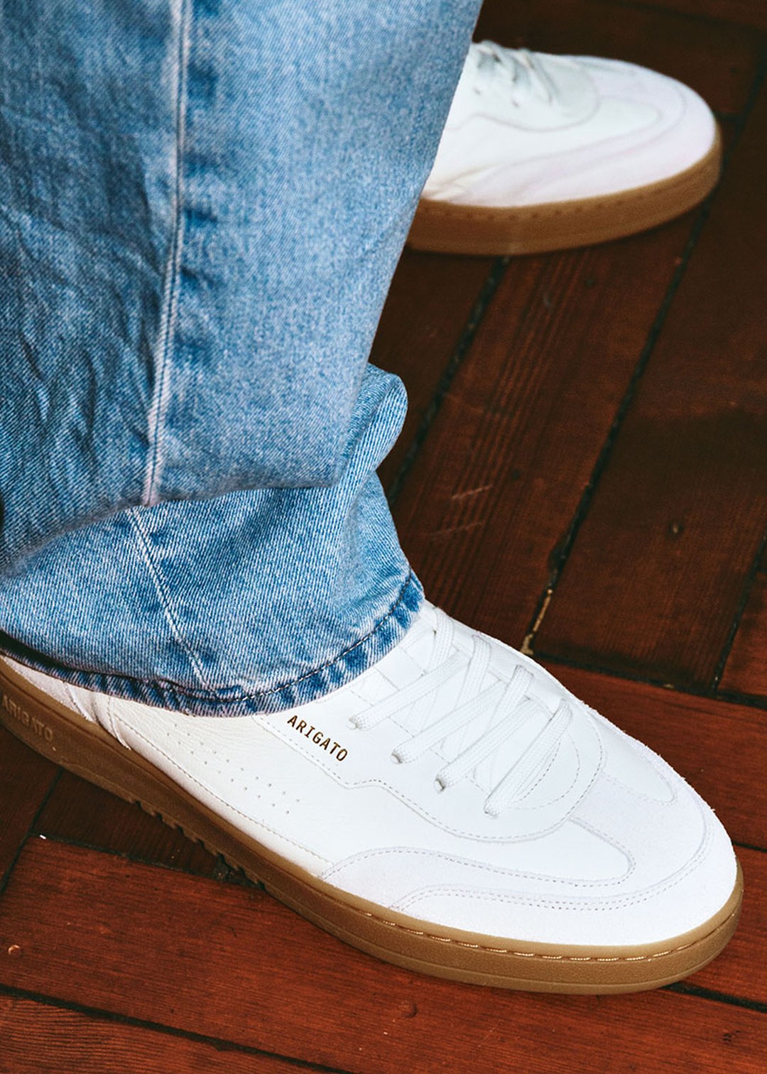 Person wearing light blue jeans and white sneakers with brown soles, standing on a wooden floor.
