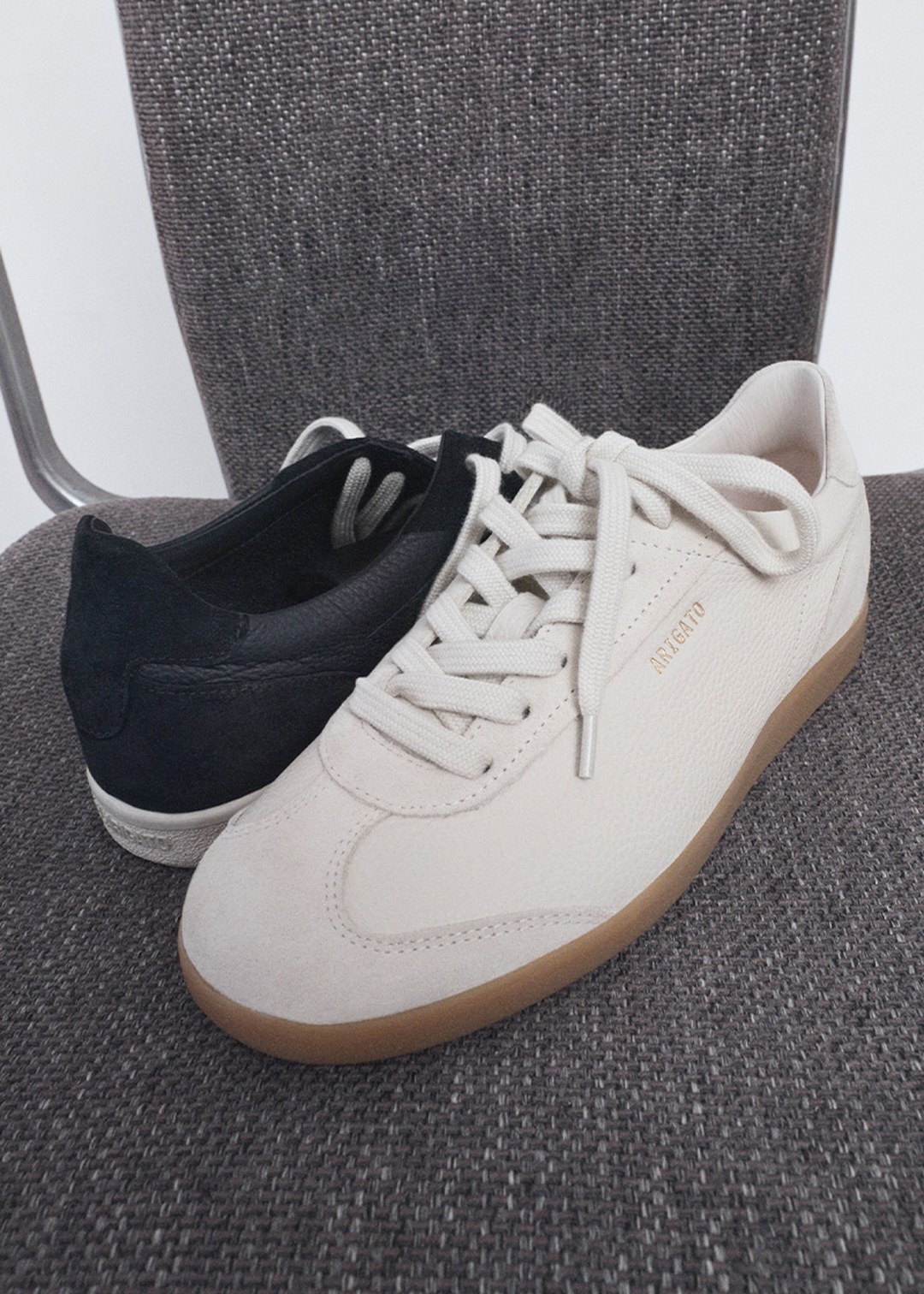 A white sneaker and a black sneaker are placed on a textured gray chair. The white sneaker has "ARGENTO" written on the side.