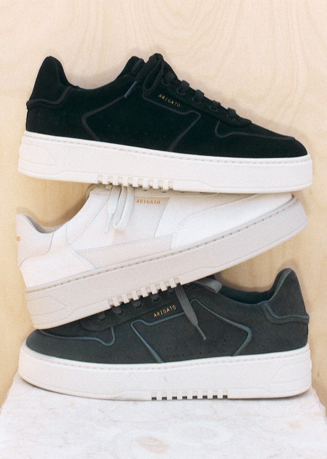 Three stacked sneakers in black, white, and gray with thick white soles, set against a light wood background.