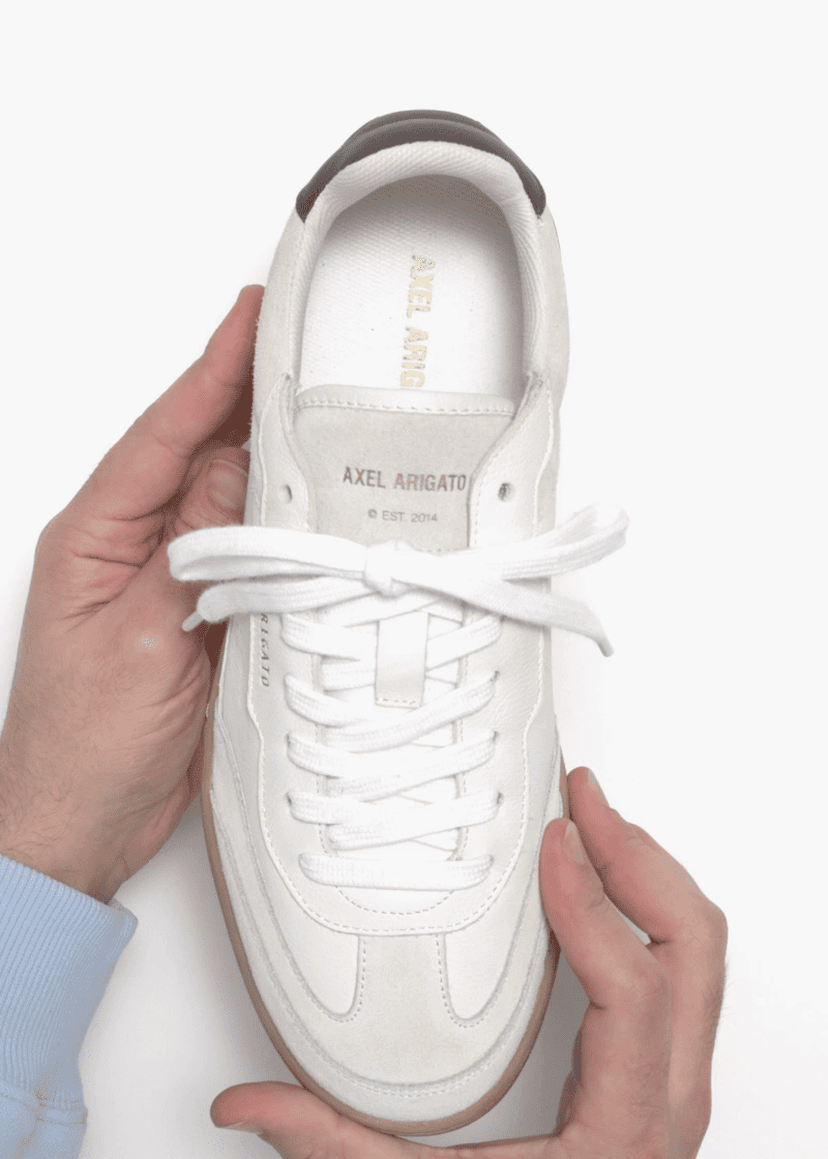 Hands holding a white Axel Arigato sneaker, showcasing its design with laces, branding, and a light brown sole, on a white background.