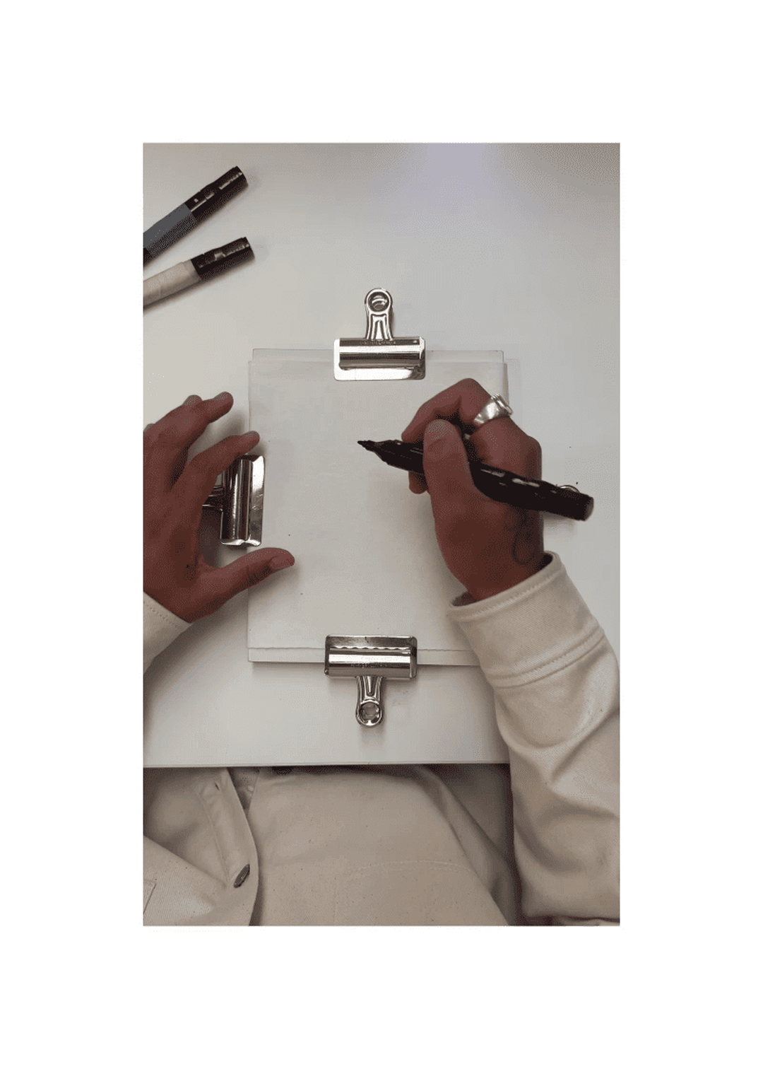 Person in white shirt drawing on paper with black marker, using a clipboard. Two markers are on the table. Hands are visible, with one wearing a ring.