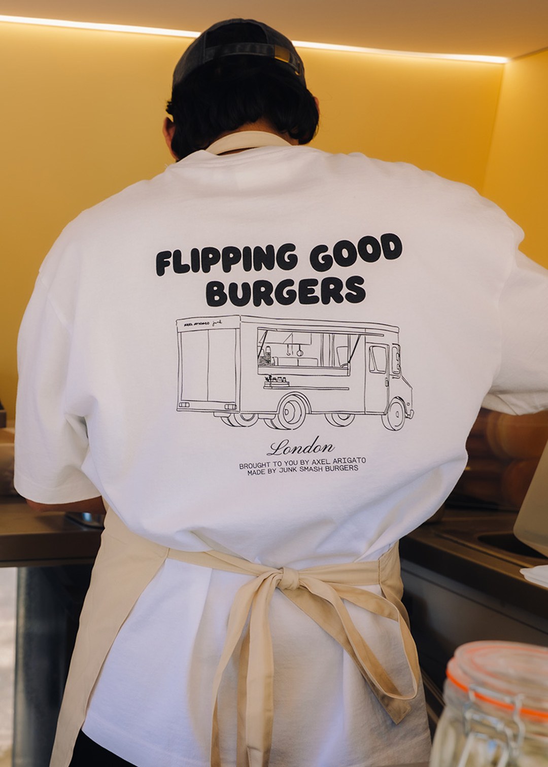 Person wearing a white T-shirt with "FLIPPING GOOD BURGERS" and a food truck illustration, standing in a kitchen with an apron.