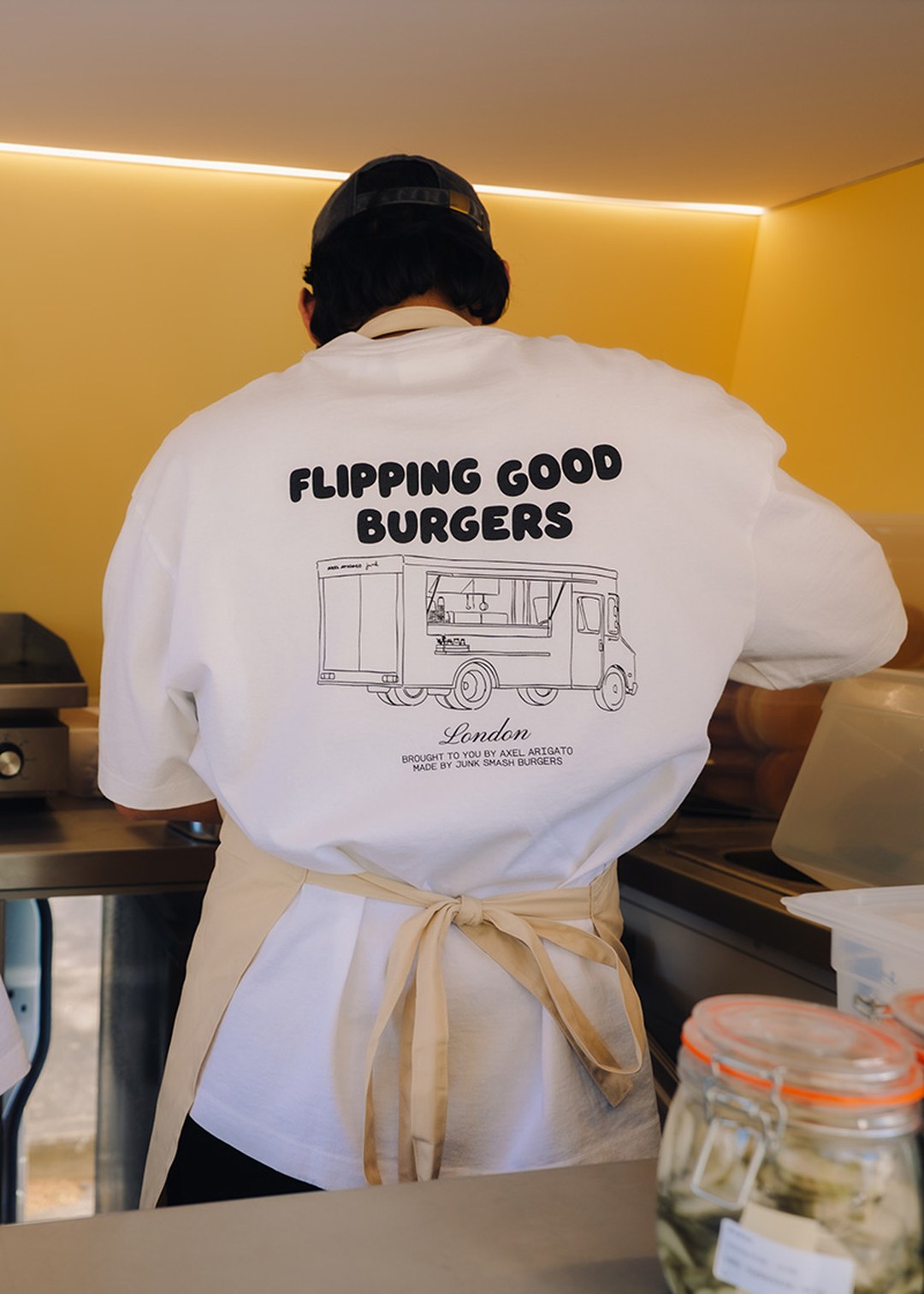 Person cooking in a kitchen wearing a shirt with "FLIPPING GOOD BURGERS" and a food truck illustration, set in a warm, well-lit space.
