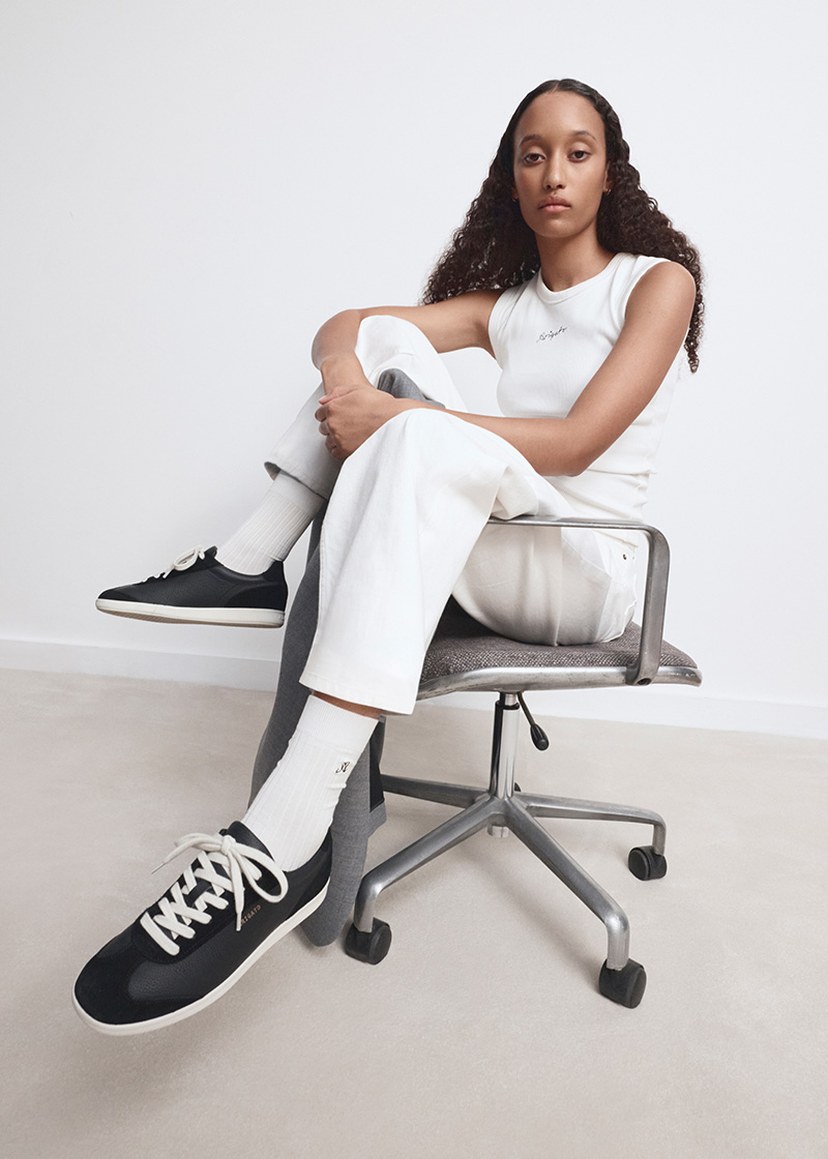 Person with long hair, in a white tank top and pants, sitting on a chair, wearing black sneakers with white laces, in a minimalist setting.