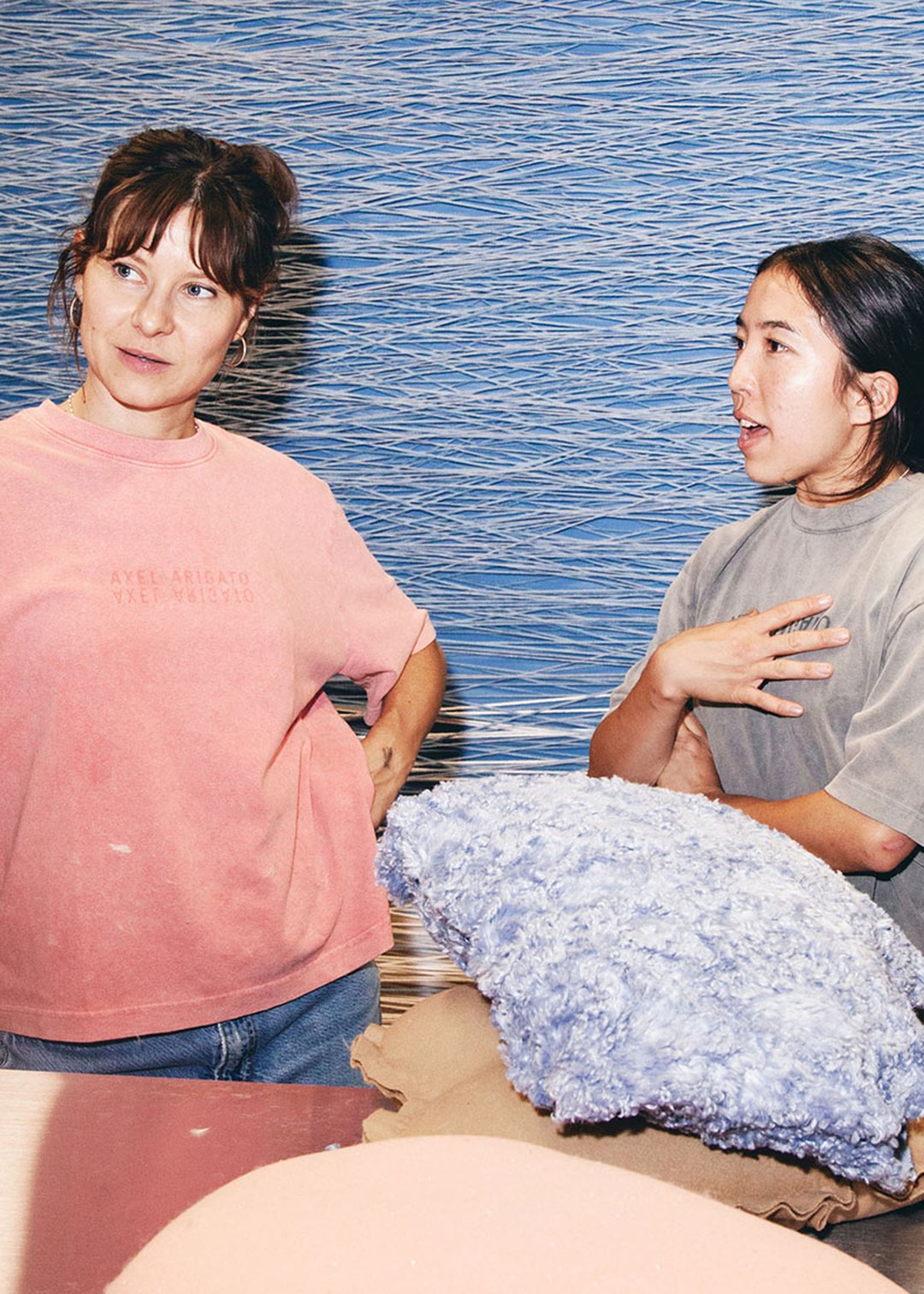 Two women in casual clothing are conversing in front of a blue textured backdrop, with a fluffy blue cushion on a table between them.