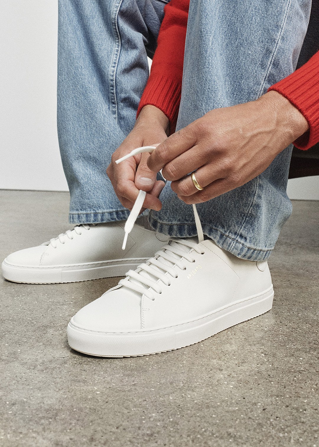 Person tying white sneakers while wearing blue jeans and a red sweater, seated on a concrete floor.