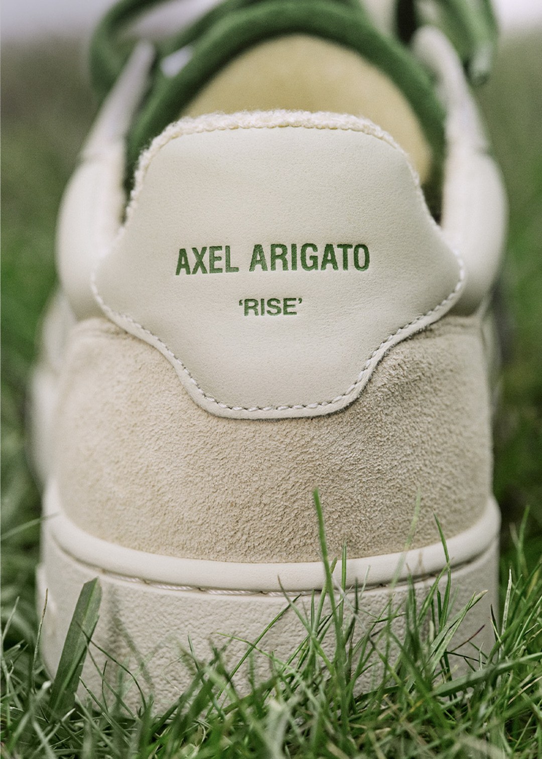 Close-up of a white Axel Arigato sneaker on grass, featuring "RISE" text on the heel in green.