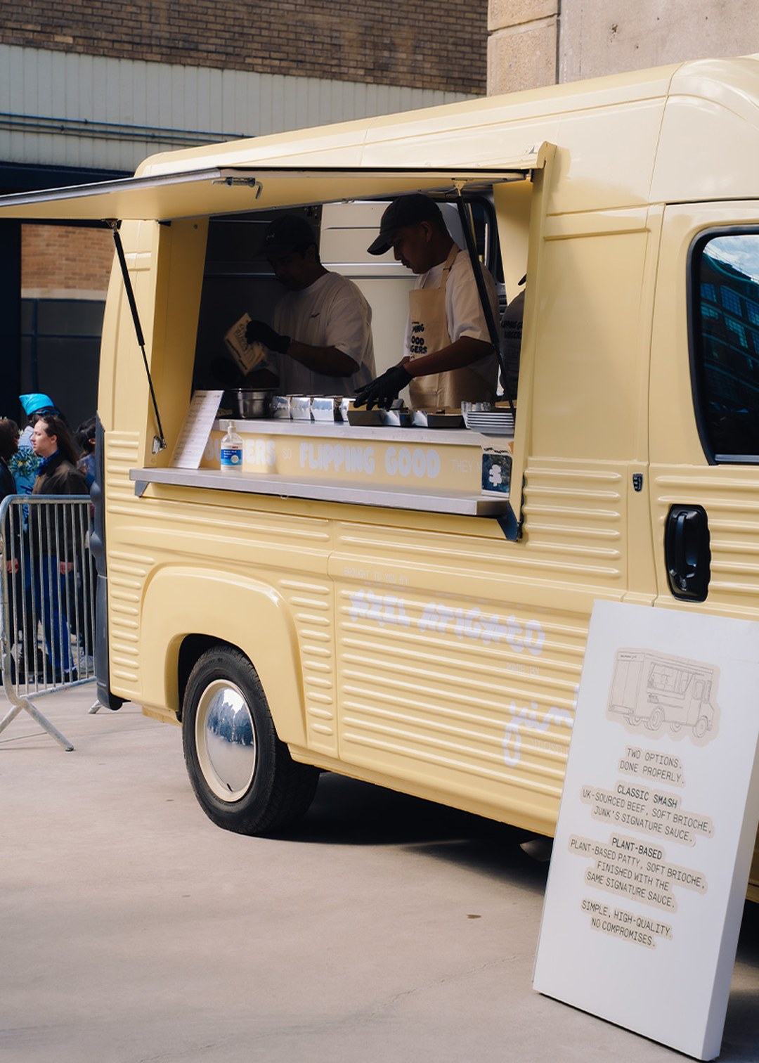 Yellow food truck with open service window, two people preparing food inside, menu board outside, and crowd in the background.