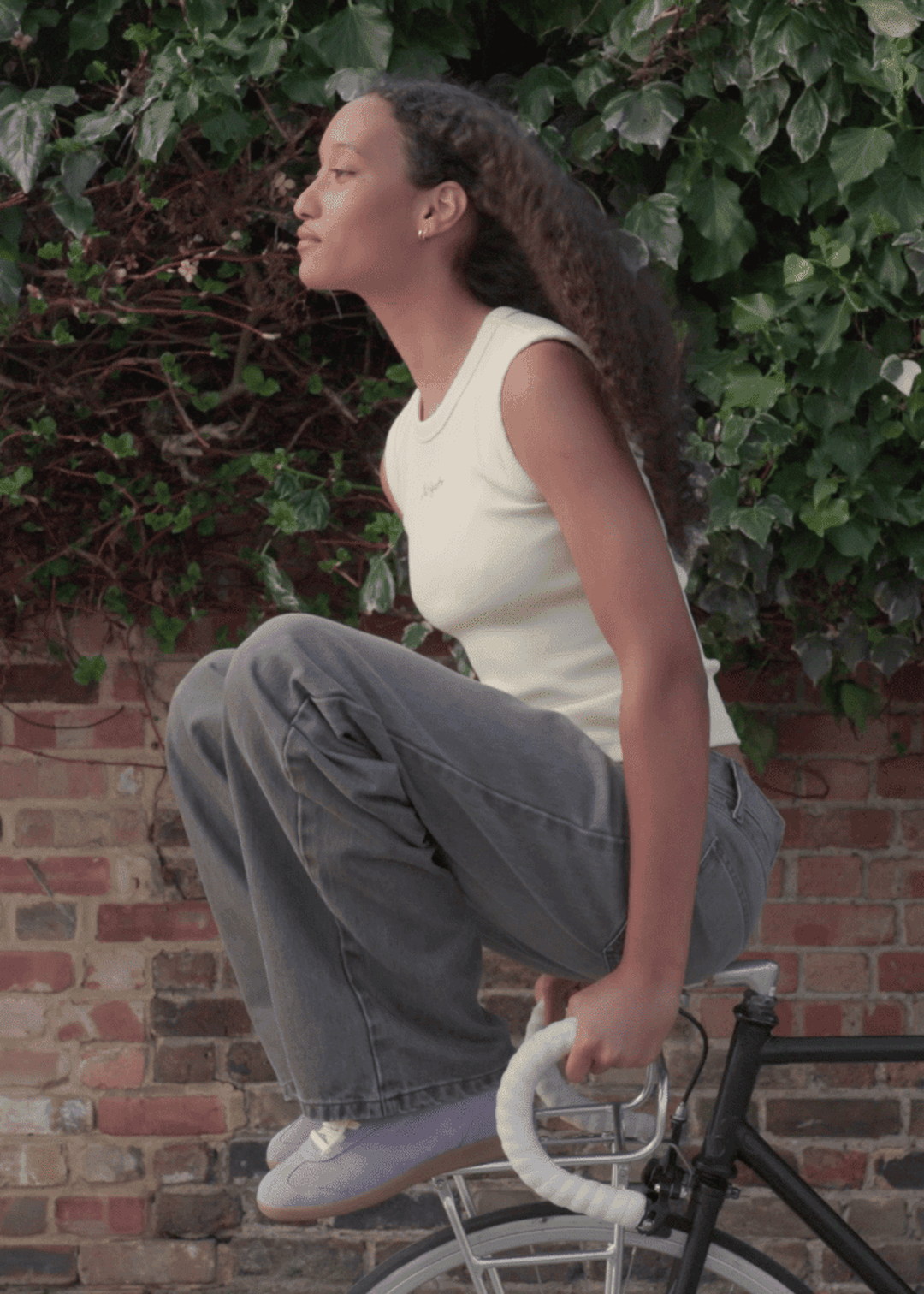Person balancing on a bike handlebar, wearing a white top and jeans, with green ivy and a brick wall in the background.