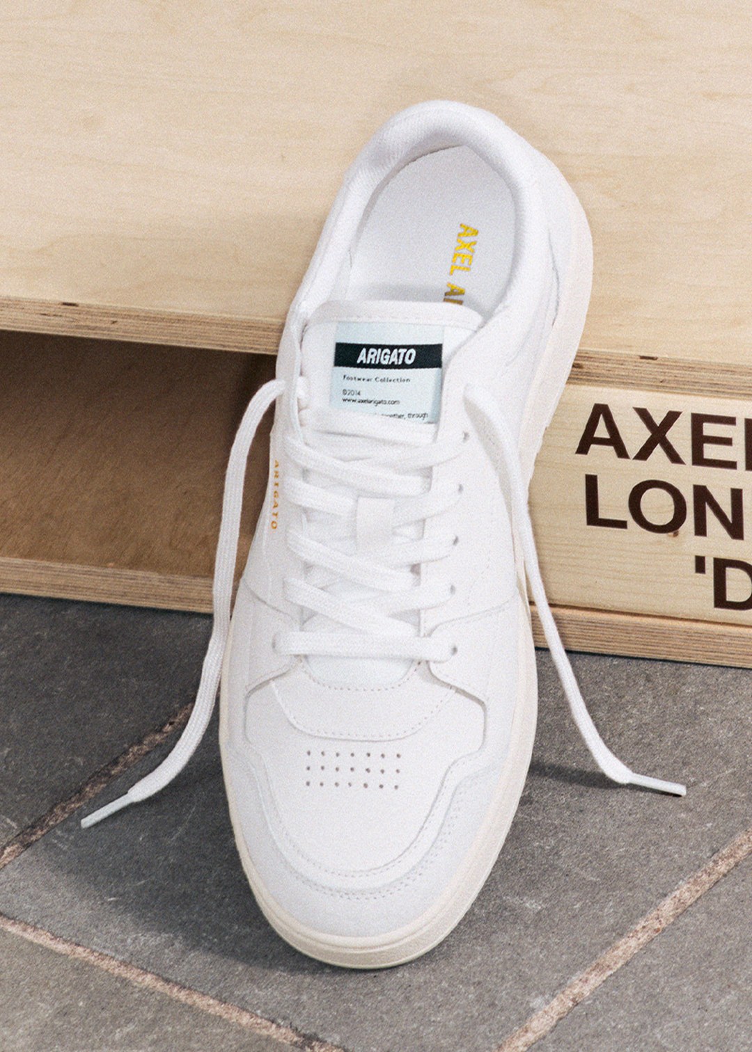White sneaker with white laces on a wooden surface, featuring a logo tag on the tongue and subtle branding on the side.