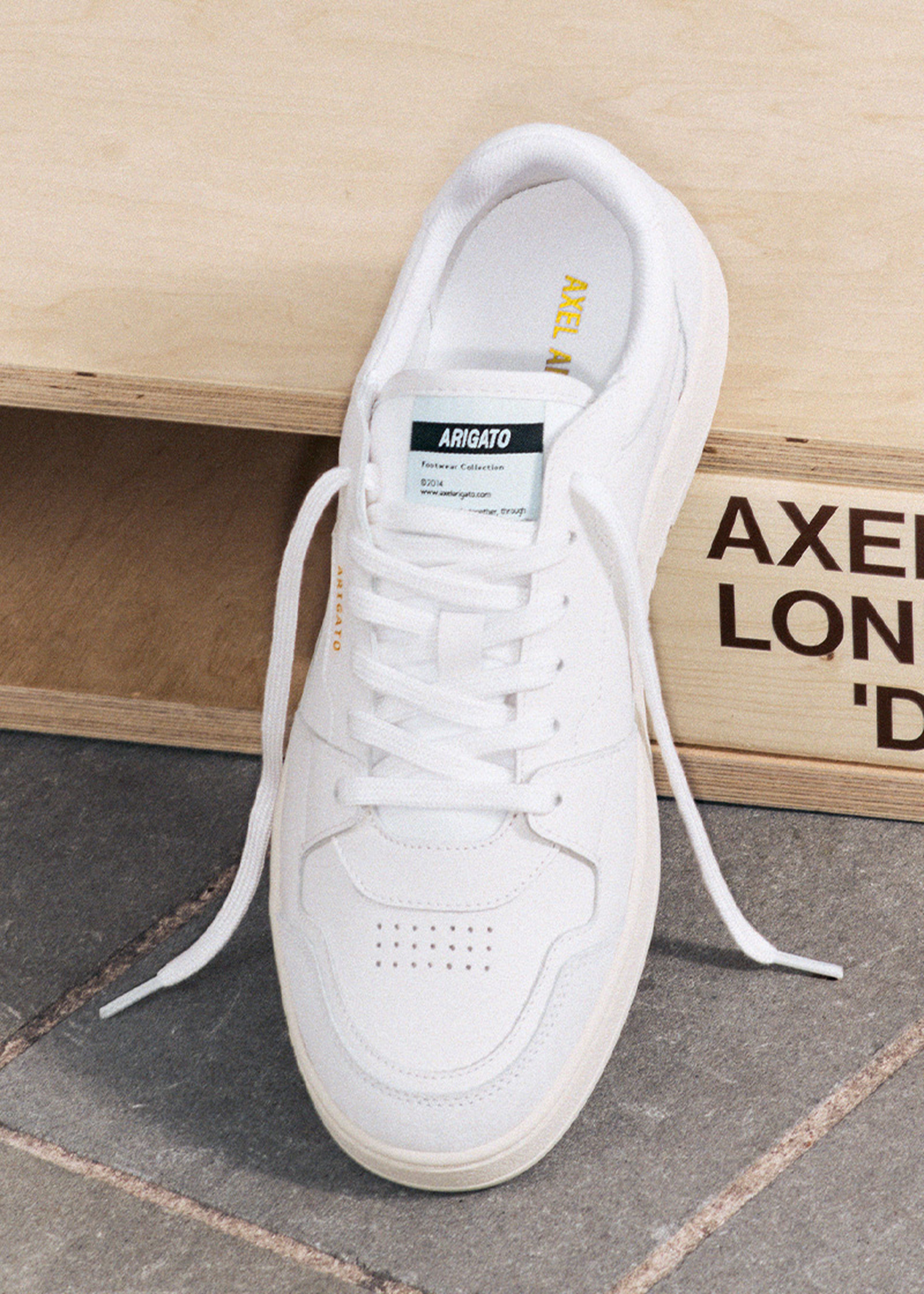 White sneaker with white laces on a wooden surface, featuring a logo tag on the tongue and subtle branding on the side.