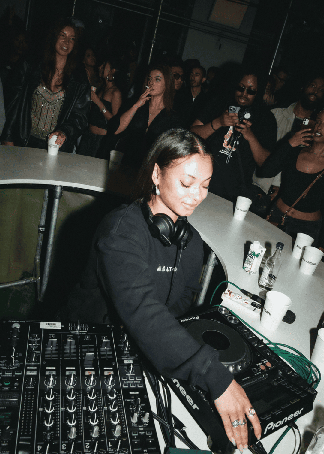 DJ performing at a lively club with a crowd watching, surrounded by DJ equipment and people holding drinks.