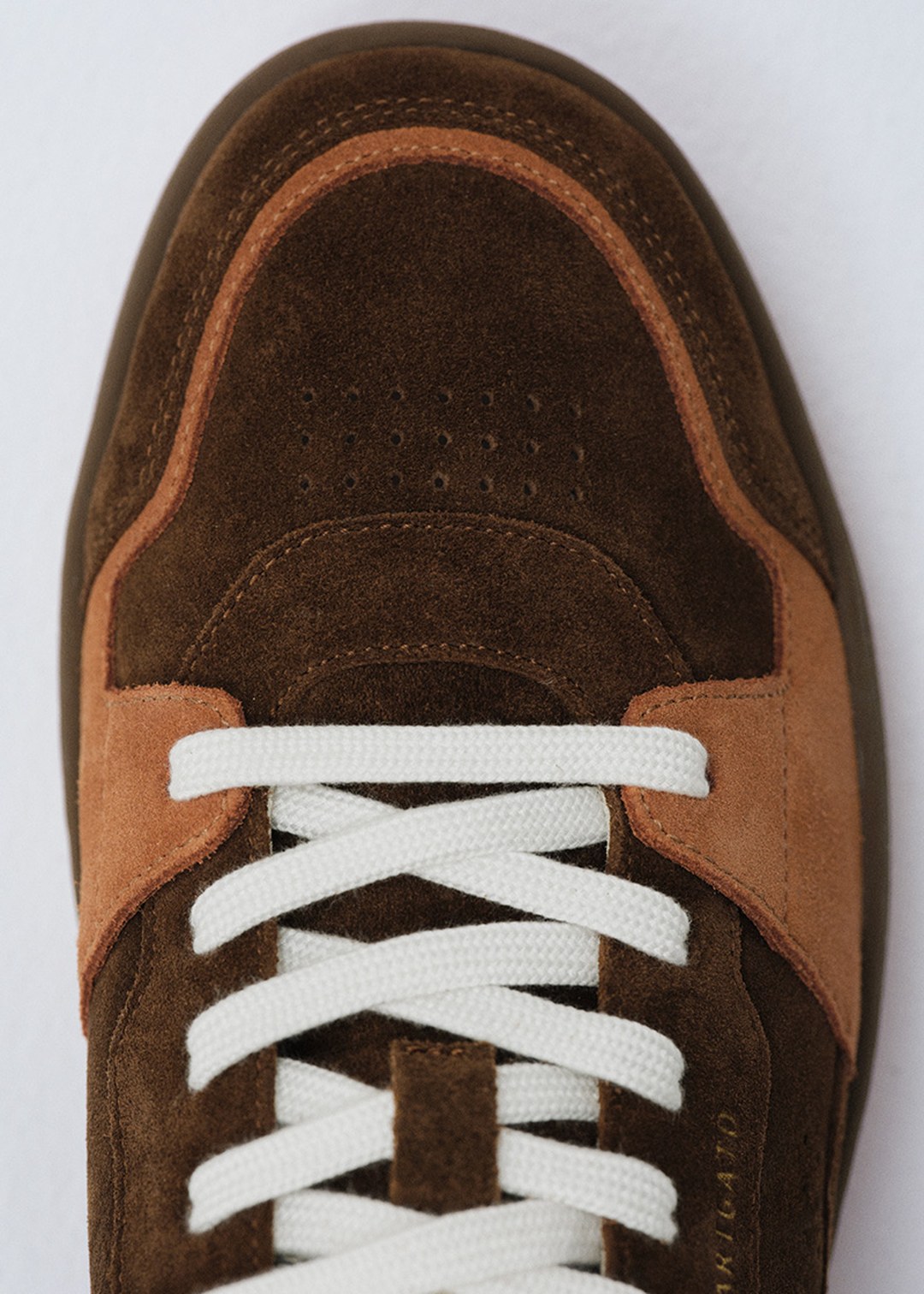 Close-up of a brown suede sneaker with light brown accents and white laces, against a plain background.
