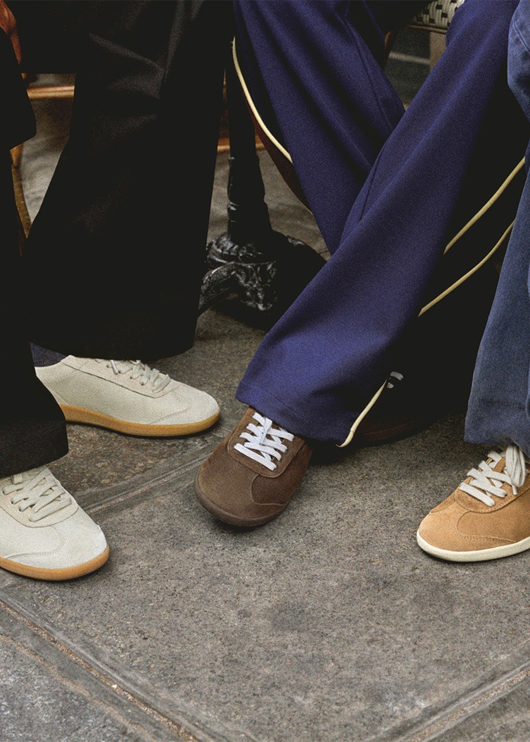 Three pairs of sneakers on a grey stone pavement: cream with gum sole, brown with white lace, and tan with cream sole.