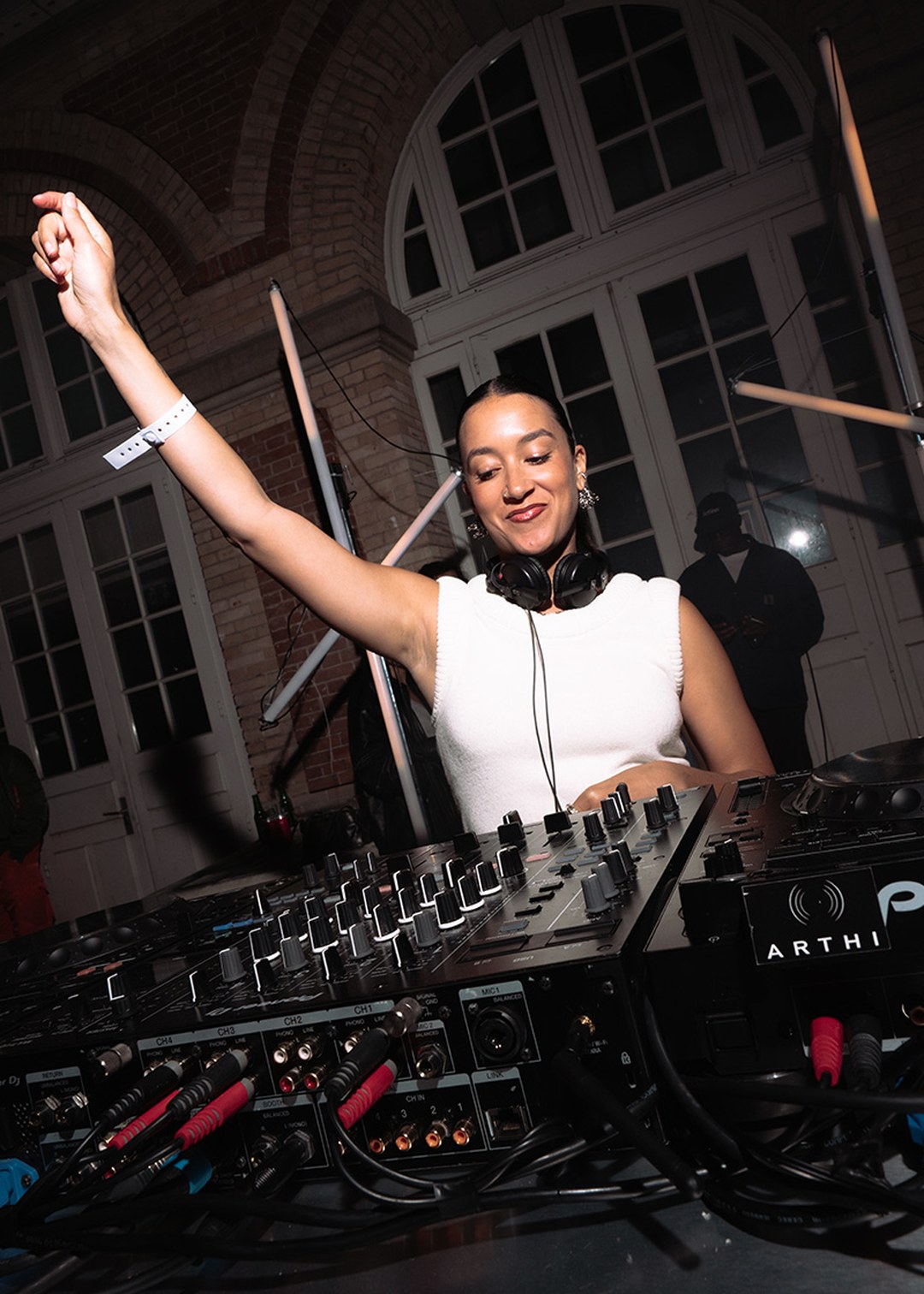 DJ in a white sleeveless top, wearing headphones, raises one arm while performing at a nightclub with a mixing console in the foreground.