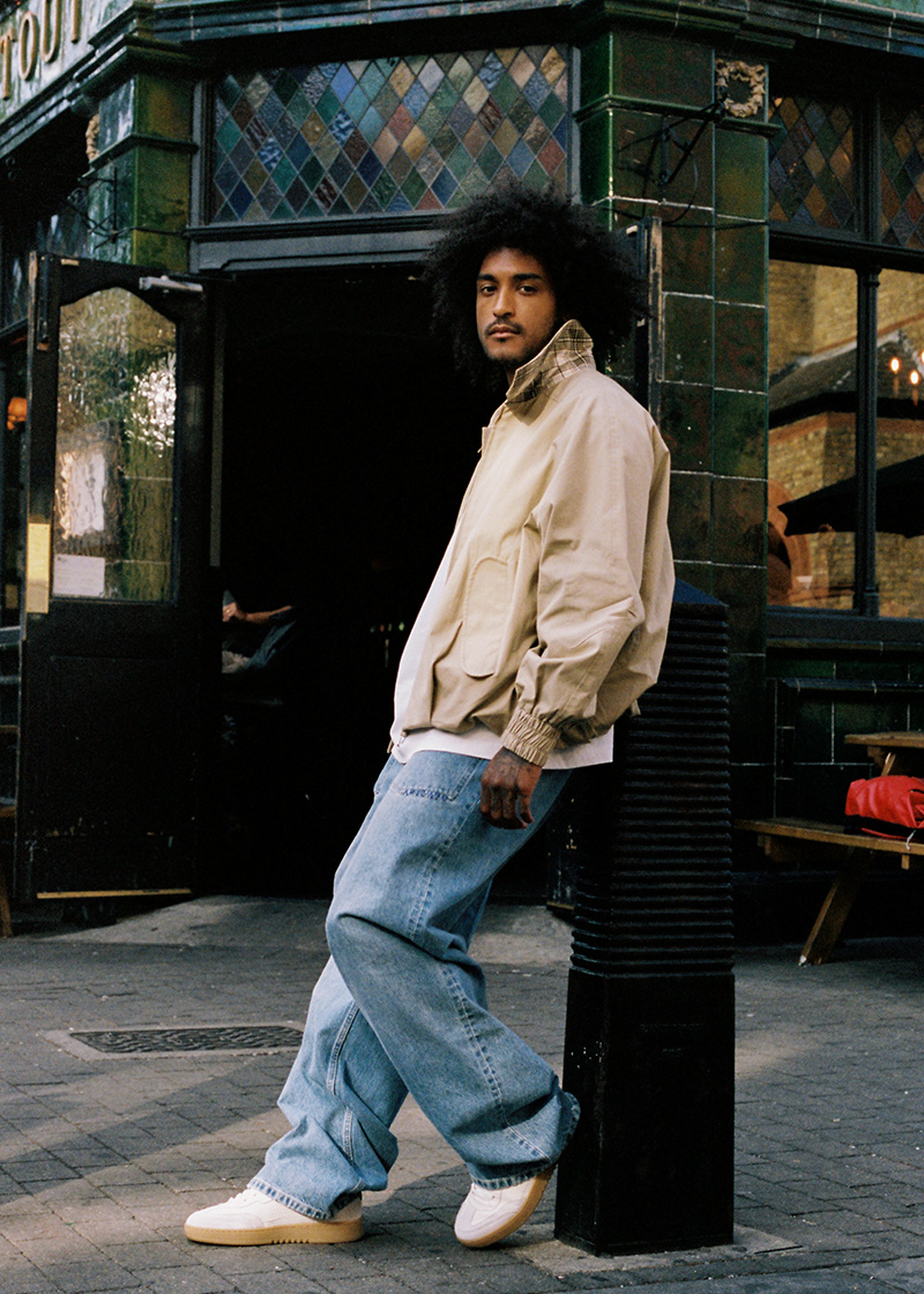 Person with curly hair wearing a light jacket leans against a green-tiled building, looking at the camera, with a relaxed pose.
