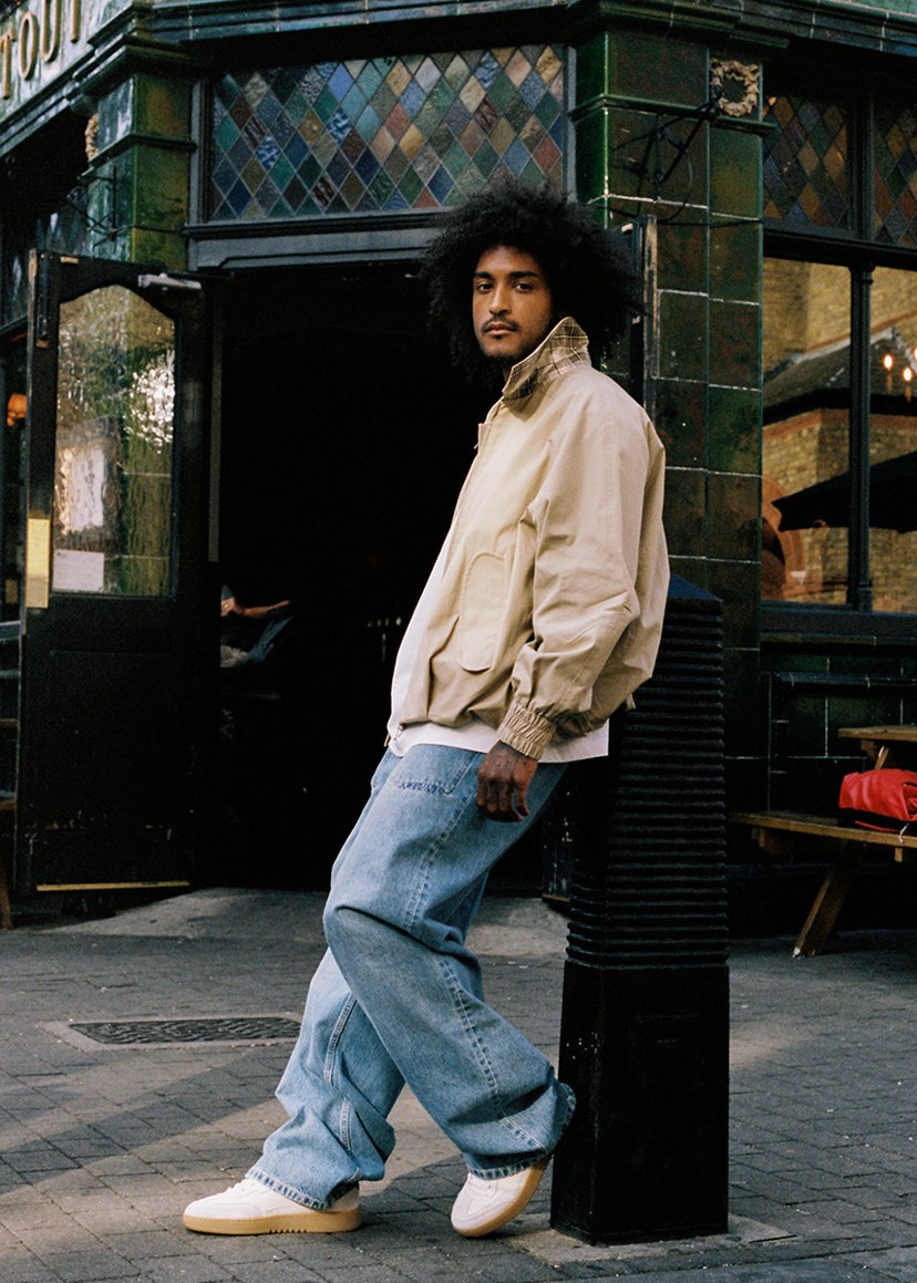Person with curly hair wearing a light jacket leans against a green-tiled building, looking at the camera, with a relaxed pose.