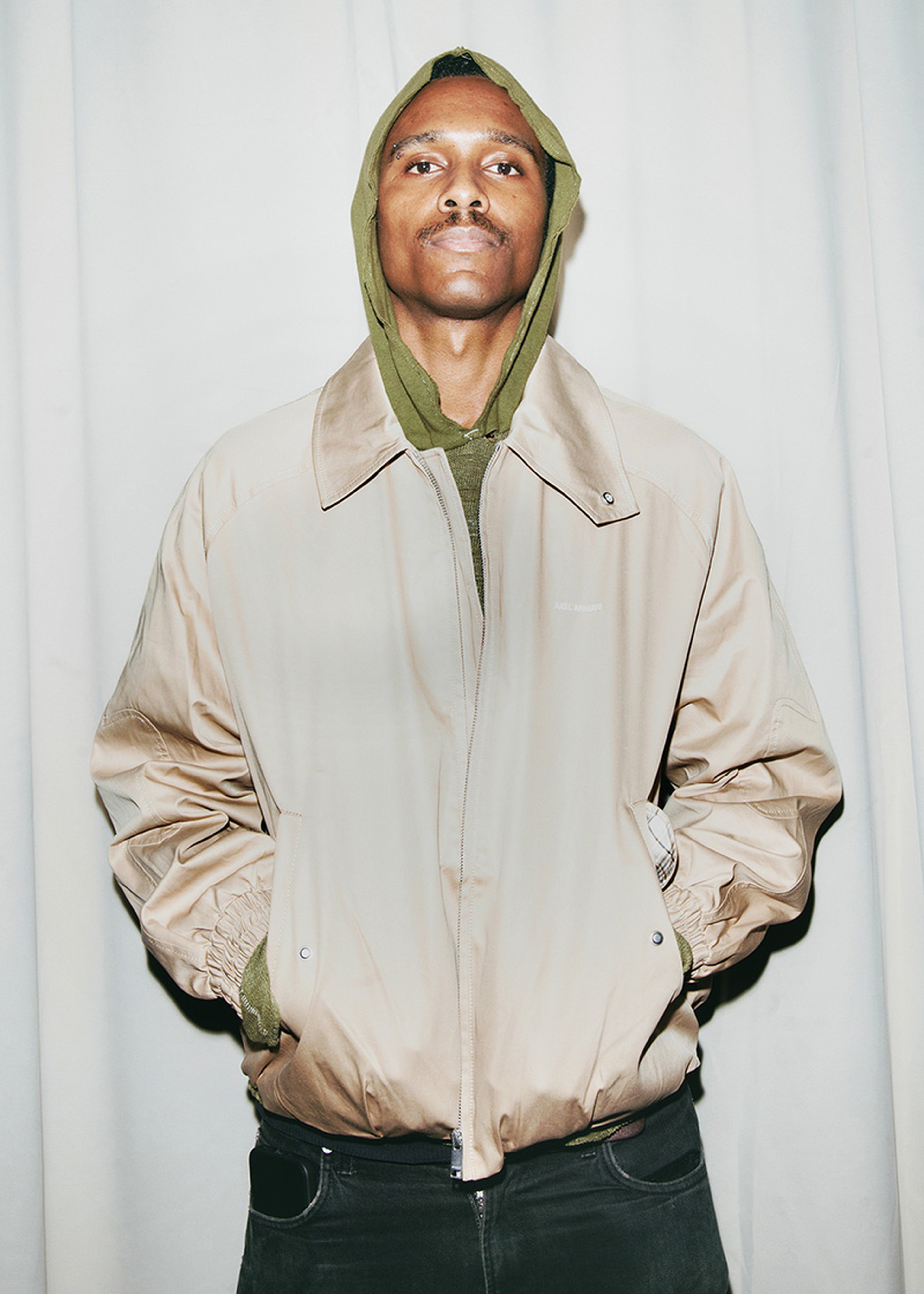 A person wearing a beige jacket over a green hoodie, with hands in pockets, standing against a plain white curtain backdrop.