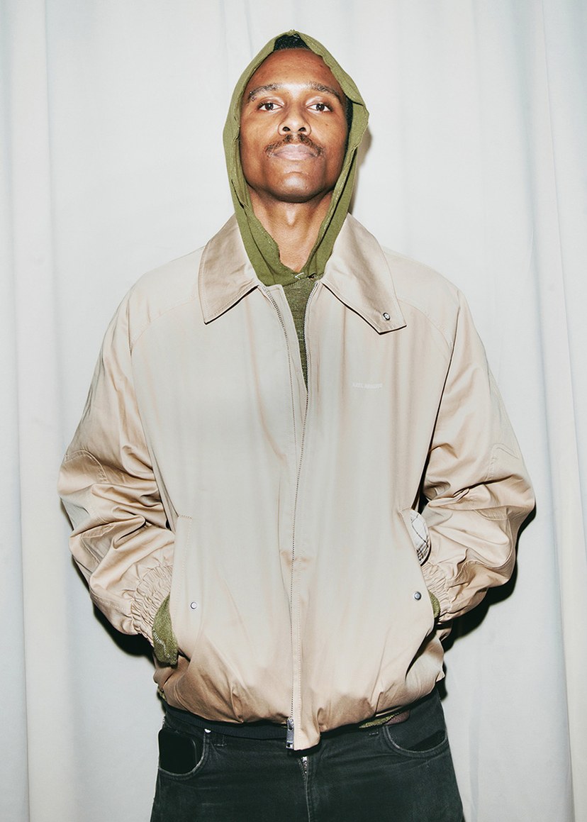 A person wearing a beige jacket over a green hoodie, with hands in pockets, standing against a plain white curtain backdrop.