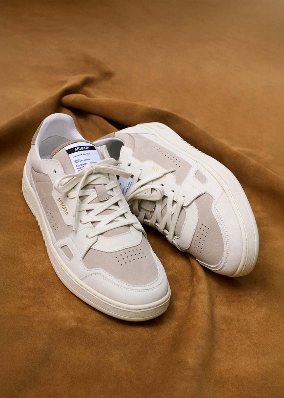 Beige and white sneakers with a minimal design, labeled "AMBUSH" on the side, placed on a soft brown fabric background.