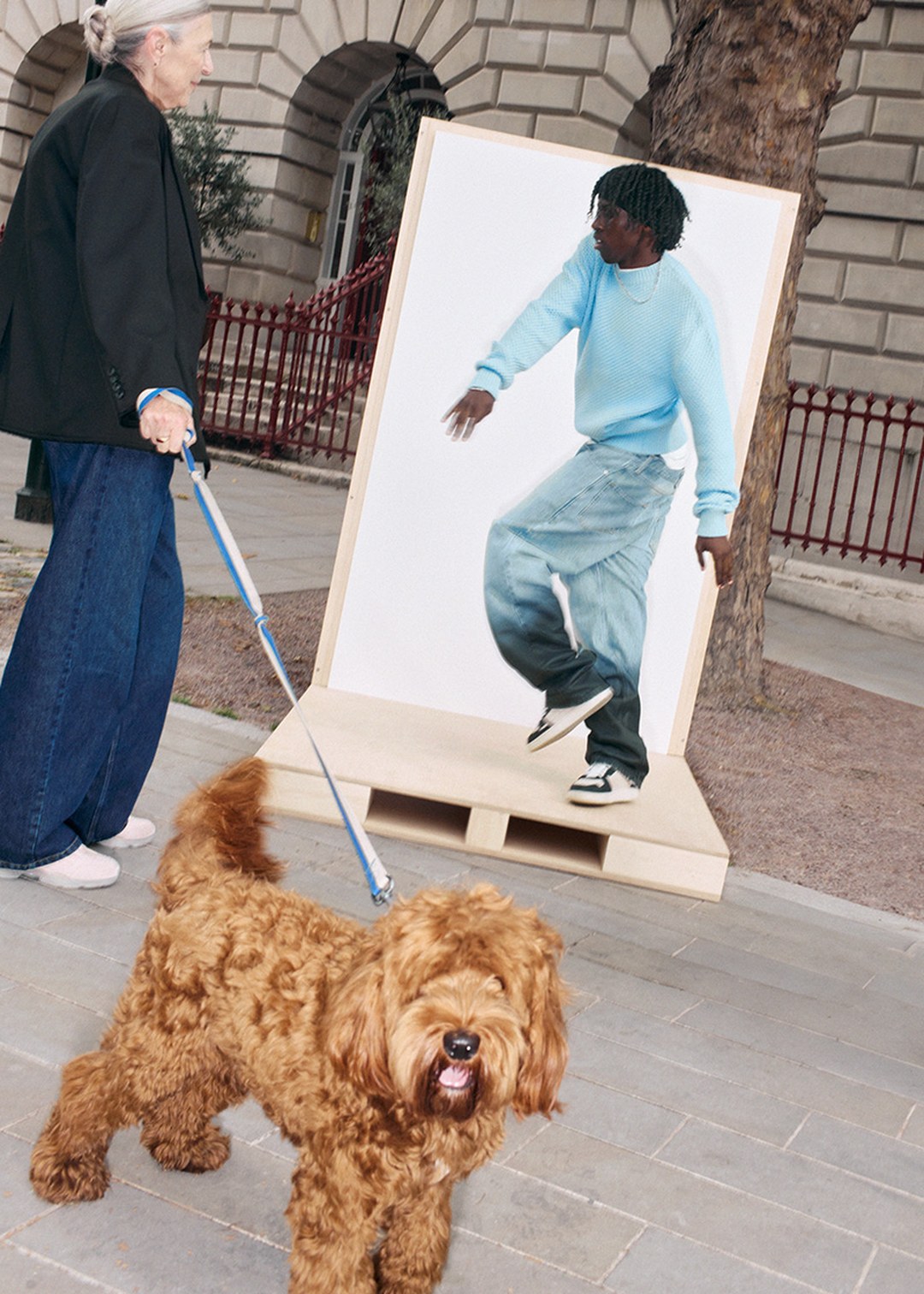 Woman walking a fluffy brown dog past a large poster of a person in a blue sweater and jeans on a city sidewalk.