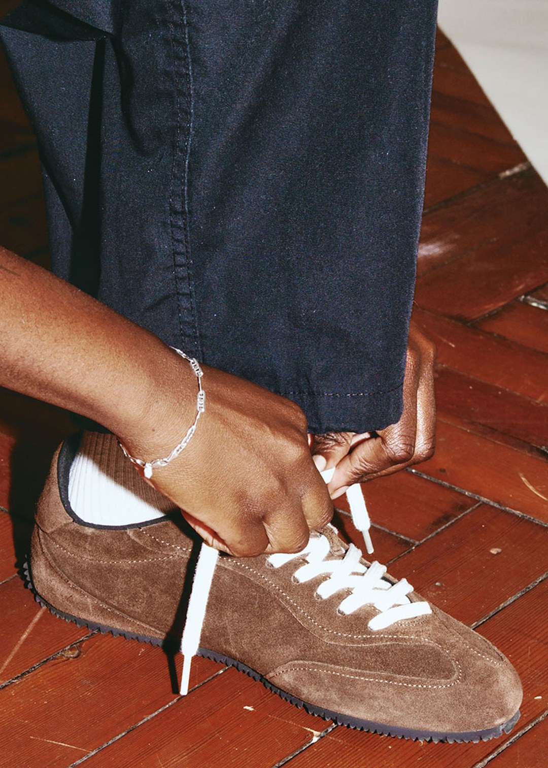 Person tying the laces of a brown suede sneaker over white socks and dark pants on a wooden floor.
