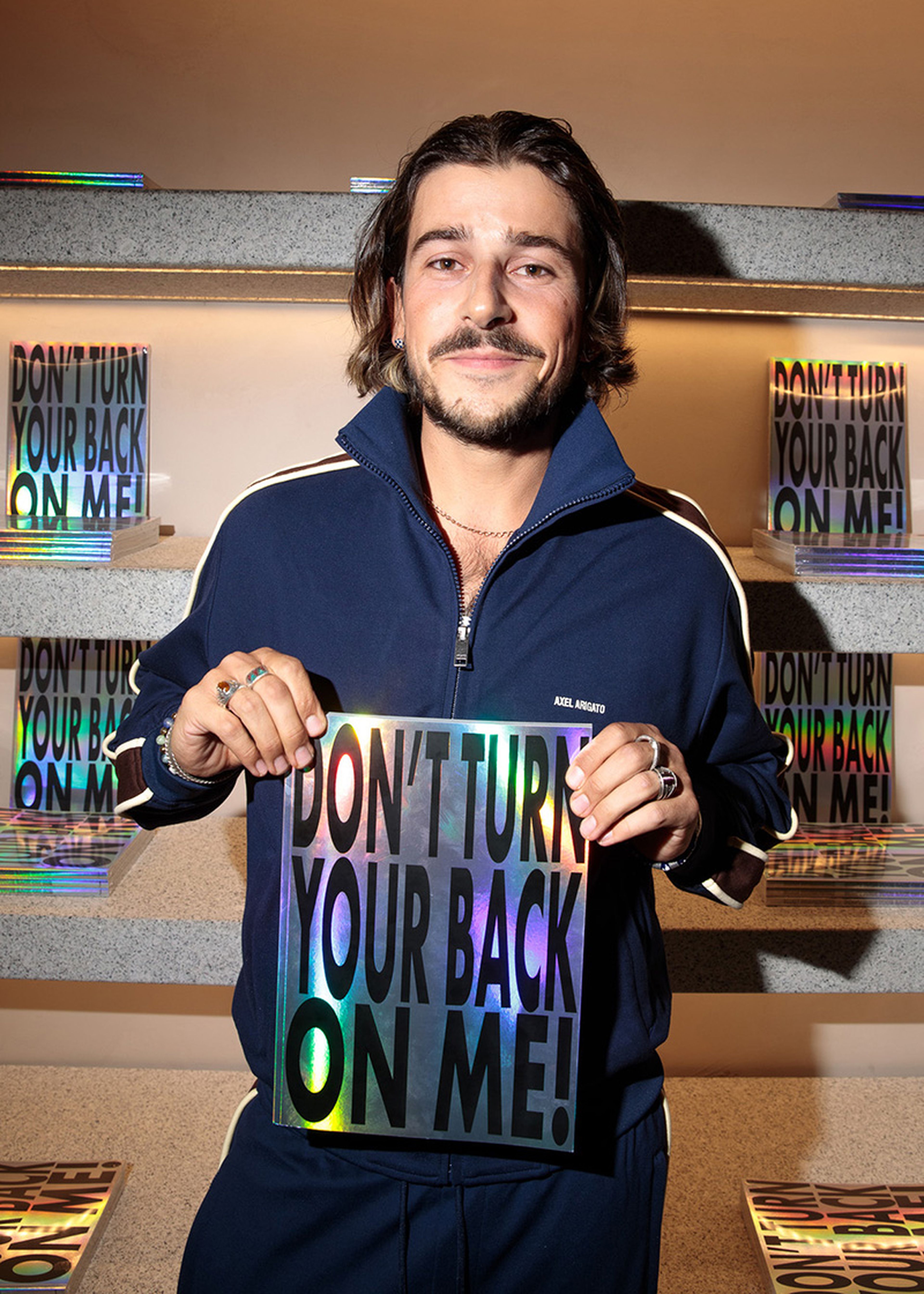 A man in a tracksuit holds a reflective sign that reads "DON'T TURN YOUR BACK ON ME!" in a room with similarly designed books on shelves.