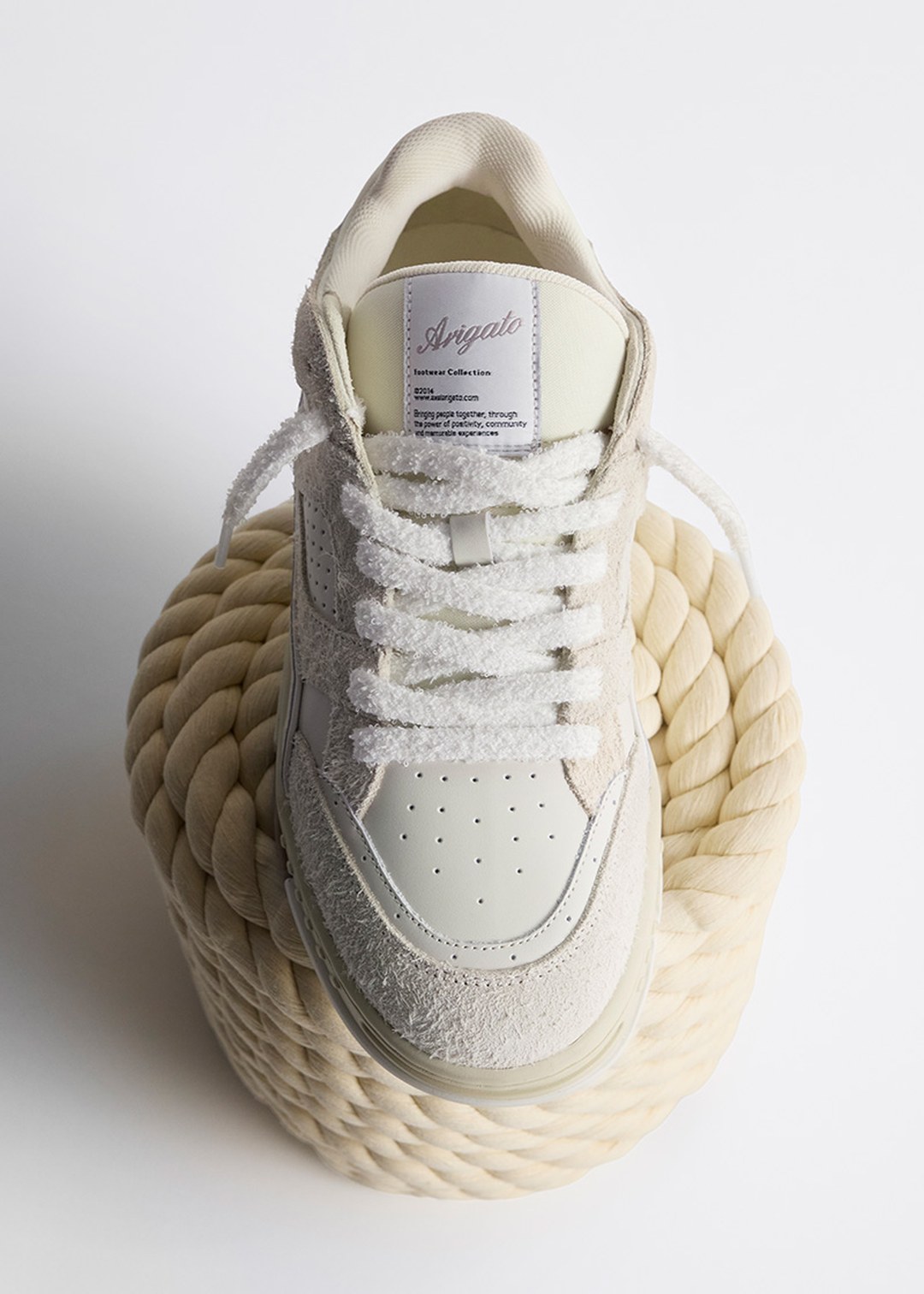 A white sneaker with textured laces is placed on a coiled rope. The sneaker features a perforated toe box and a label on the tongue.