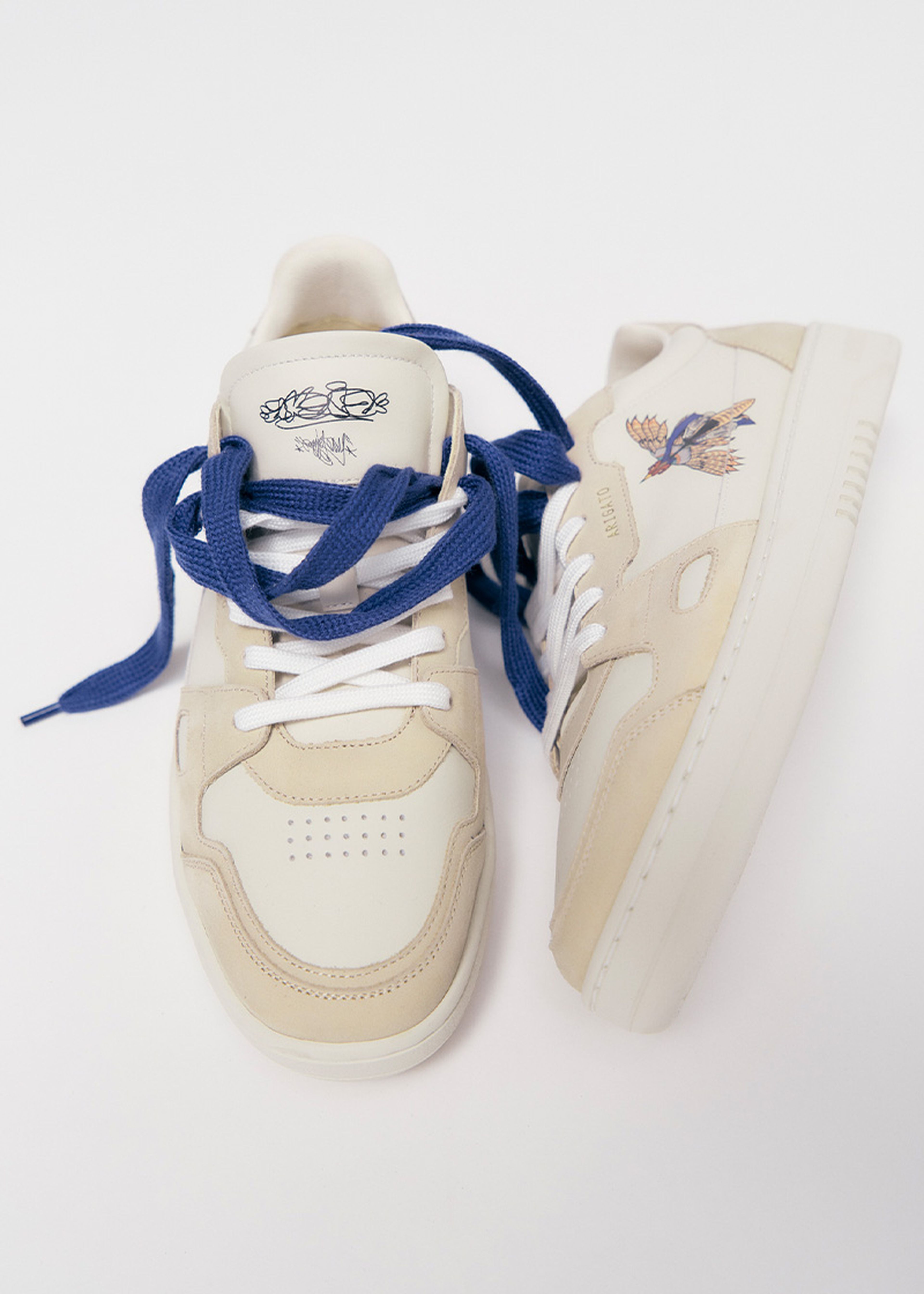 Beige sneakers with navy blue laces, one featuring an embroidered bird design on the side, against a plain white background.