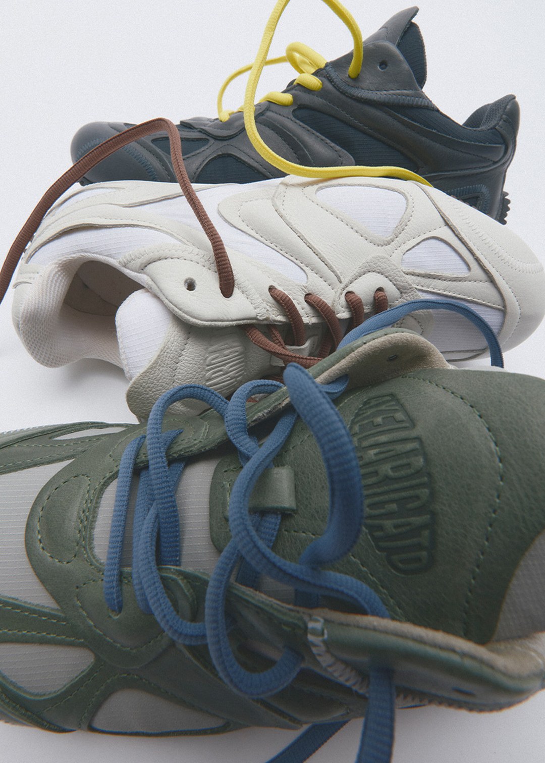 Three sneakers with intertwined colorful laces: dark green, white, and gray. Each shows unique textures and overlapping designs.