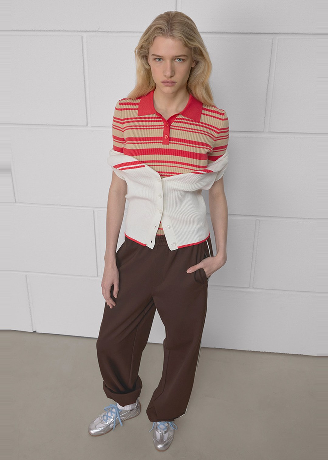 Person with long blonde hair wearing a red striped top, white cardigan, brown pants, and silver shoes, standing against a white brick wall.