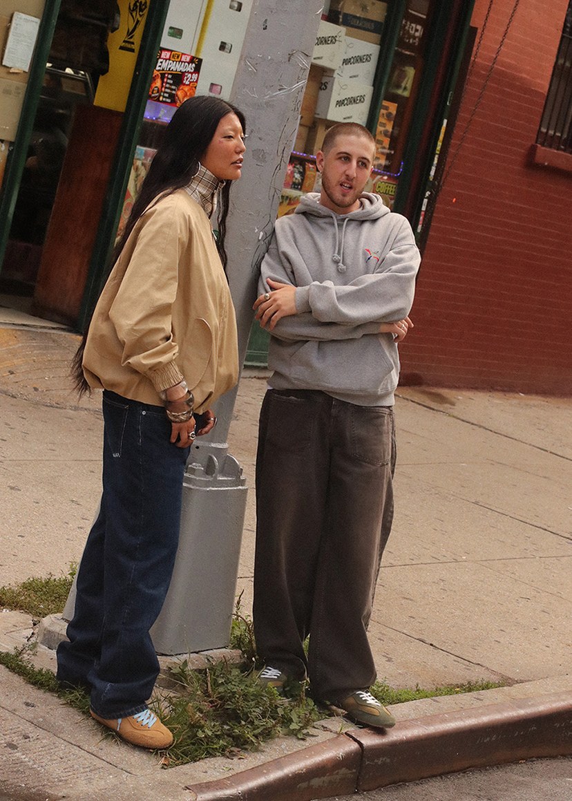 Two people stand on a city sidewalk, with one wearing a beige jacket and jeans, the other in a gray hoodie and dark pants, near a brick building.