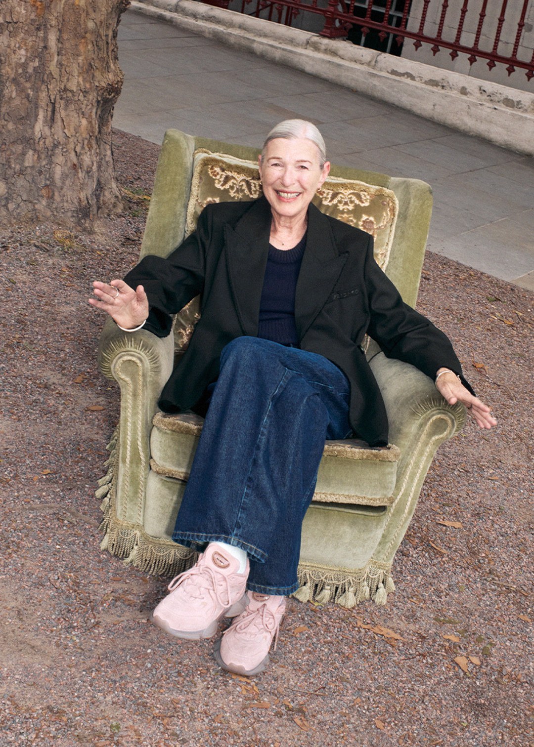 Elderly woman smiling, seated in a green armchair outdoors, wearing a black blazer, blue jeans, and pink shoes, with a tree in the background.
