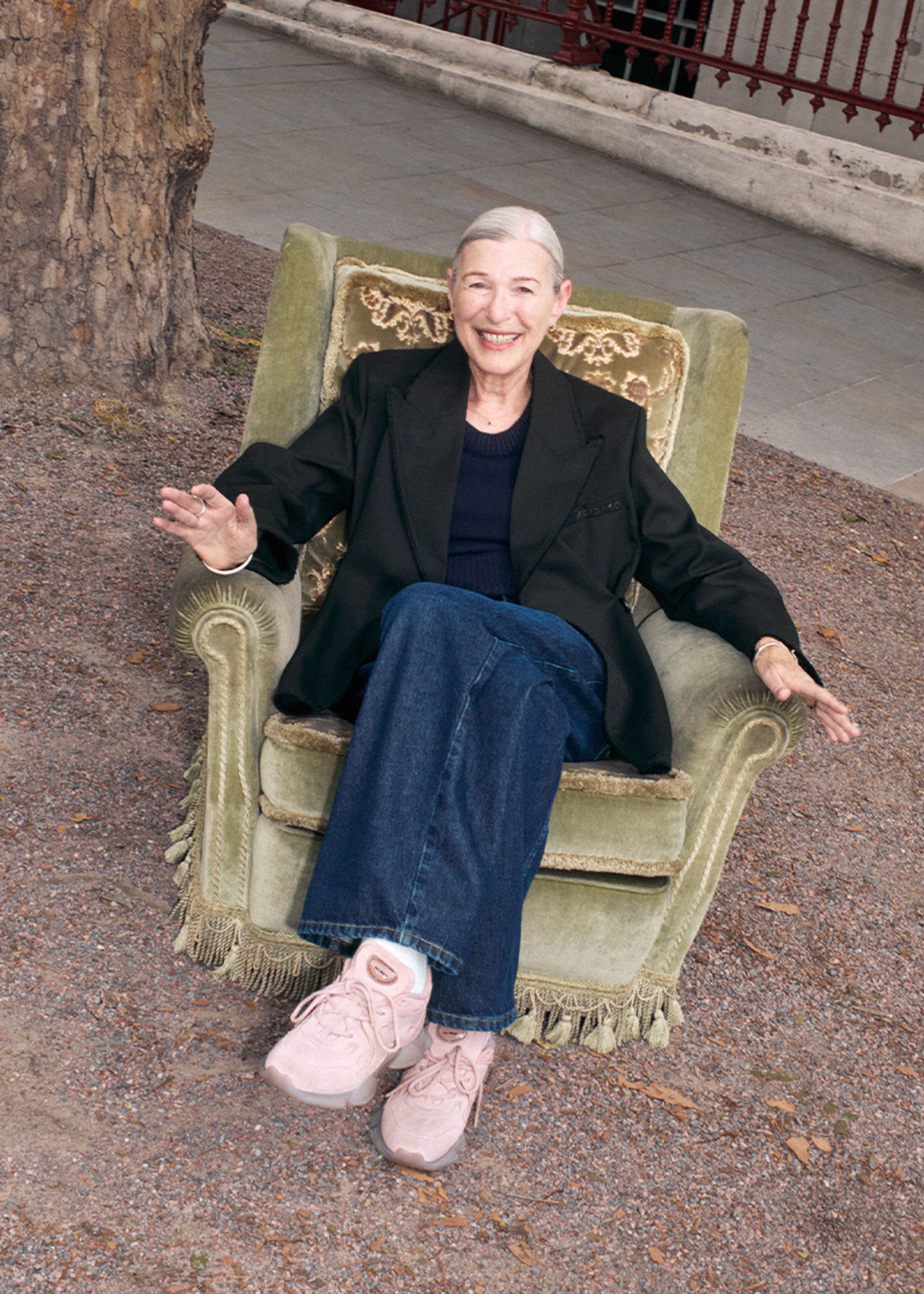 Elderly woman smiling, seated in a green armchair outdoors, wearing a black blazer, blue jeans, and pink shoes, with a tree in the background.