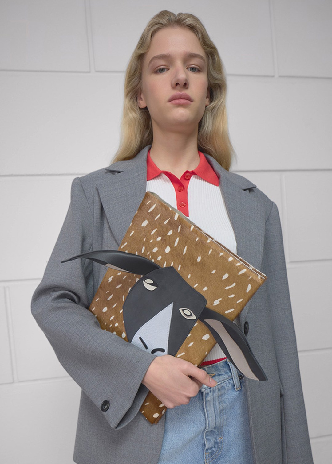A person wearing a gray blazer and red-collared shirt holds a brown folder with a stylized animal face design against a white tiled wall.