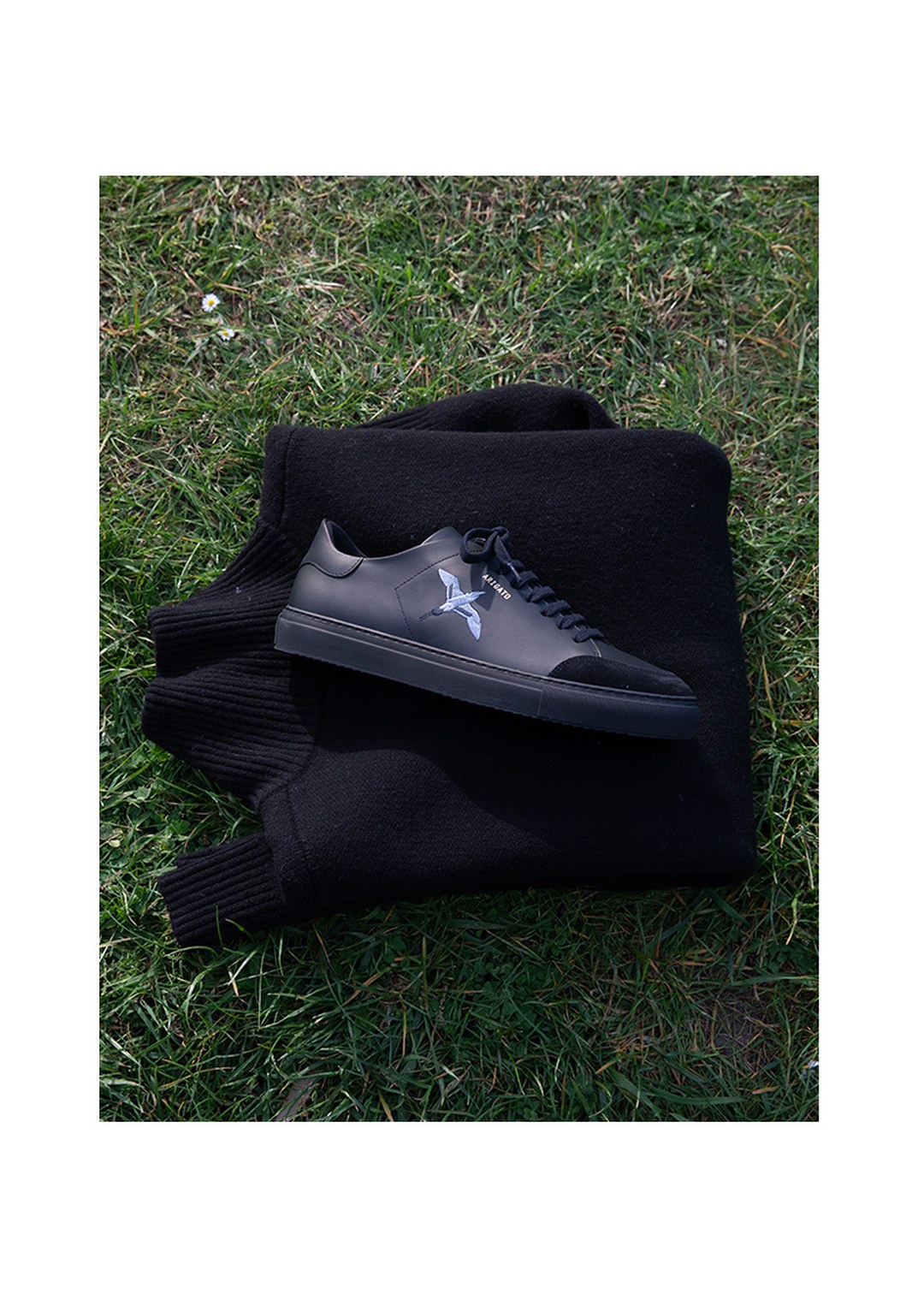Black sneaker with star logo on a folded black sweater, placed on green grass.