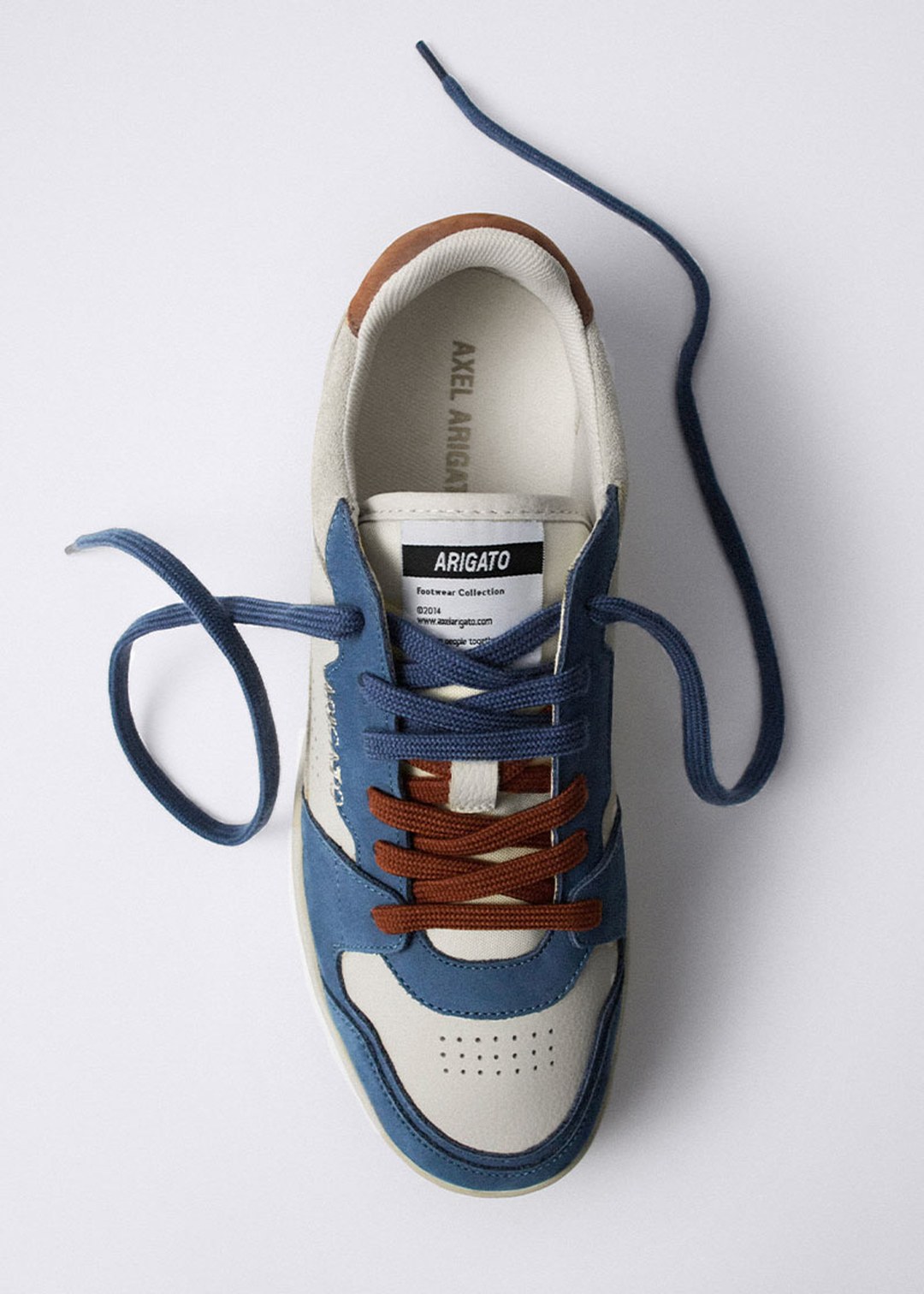 Top view of a blue and white sneaker with brown laces, featuring a label inside that reads "Arigato" and a dark blue loose lace.