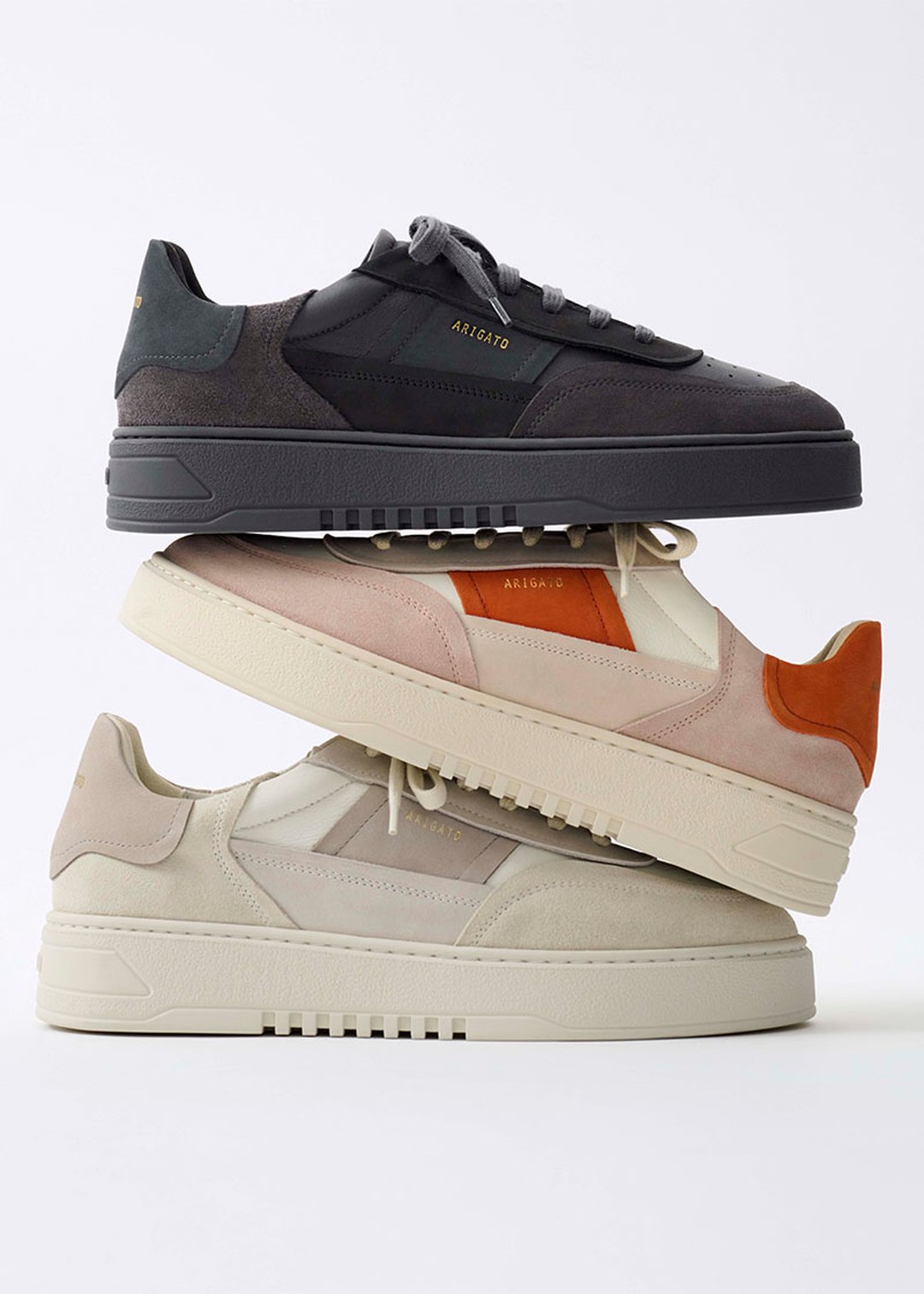 Stack of three stylish sneakers in dark gray, beige, and cream with orange accents against a plain white background.