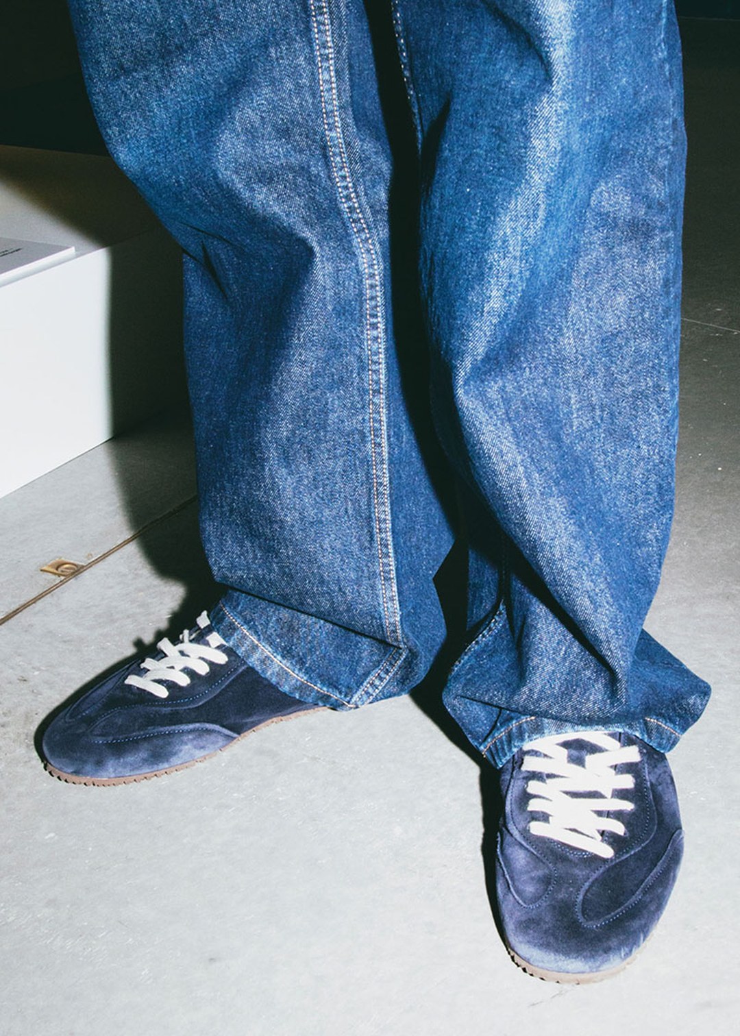 Person wearing loose blue jeans and navy sneakers with white laces, standing on a light-colored floor.